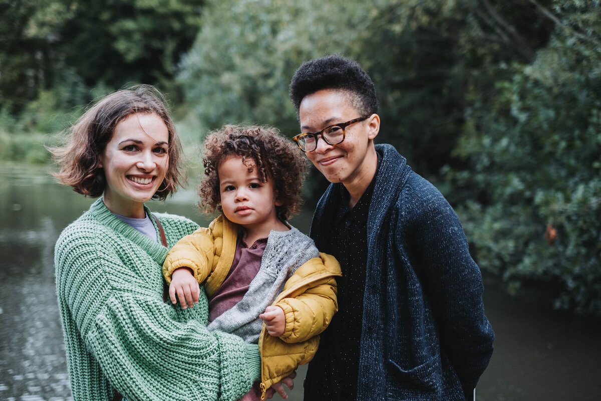 Curly haired girl is held by her mums in front of pond in Hampstead London