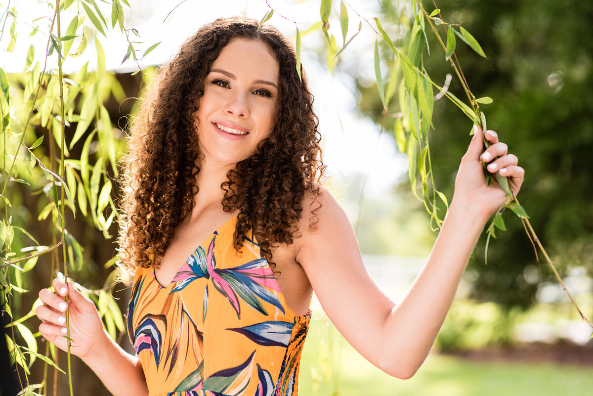 High school senior girl with naturally curly hair wearing floral dress stands in a willow tree at Amber Grove poses for her senior portraits.