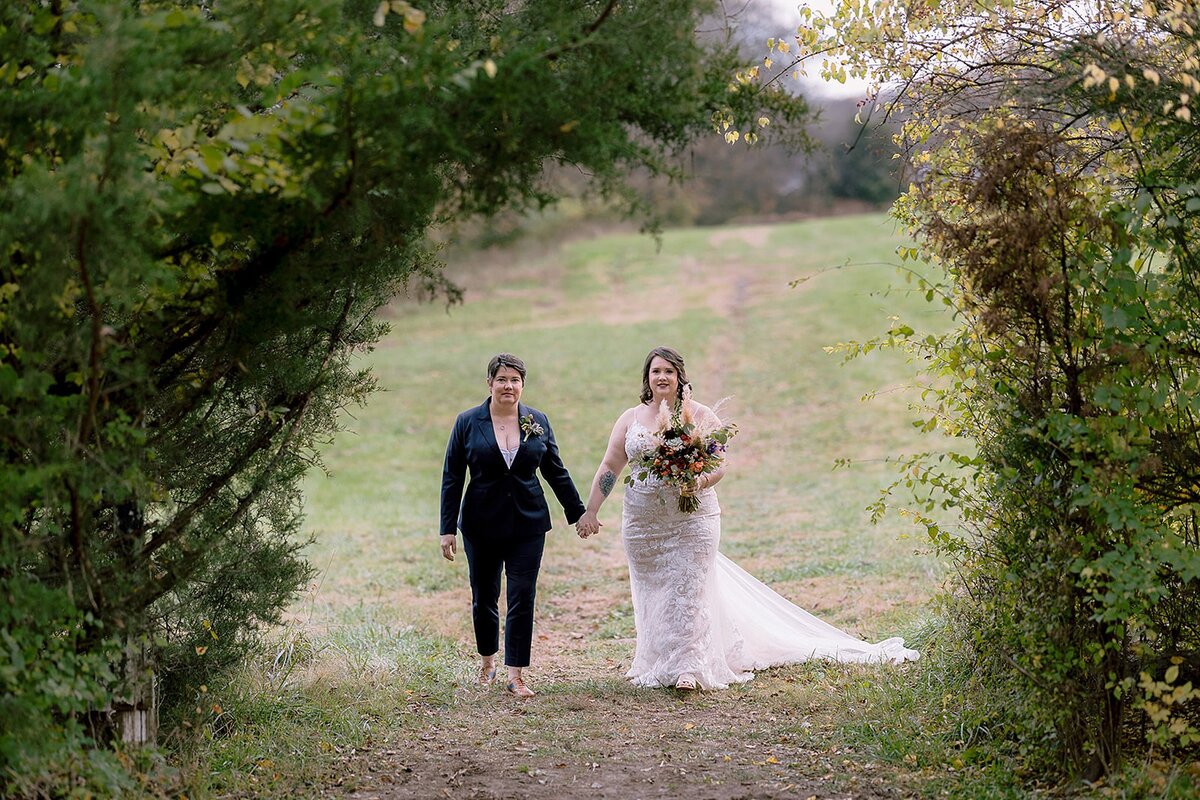A bride wearing a navy blue suit walks hand in hand with a bride wearing a fitted white lace wedding dress with a long train. They are walking through an arbor of trees in a field at Ravenswood Mansion.