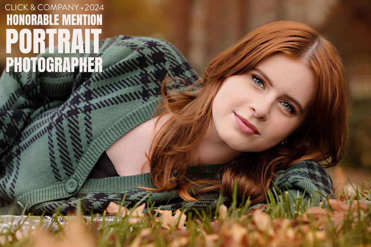 Portrait of a young lady with red hair wearing a green cardigan and laying in the grass.  There is a watermark logo in the corner saying "Click & Company 2024 Honorable Mention Portrait Photographer"