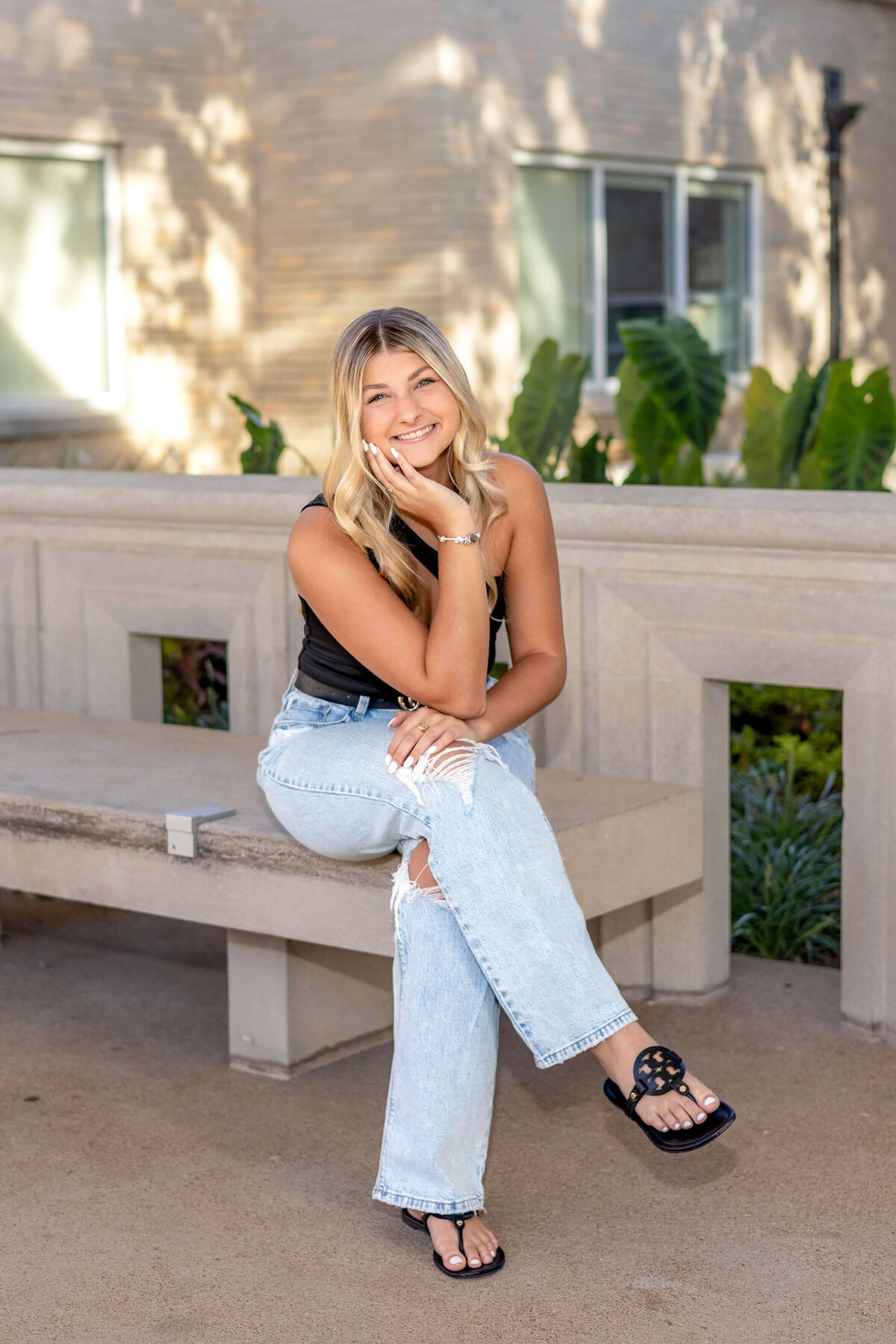 a blonde haired high school senior girl sitting  on a concrete bench with legs crossed and elbow resting on leg and hand resting under face smiling at the camera