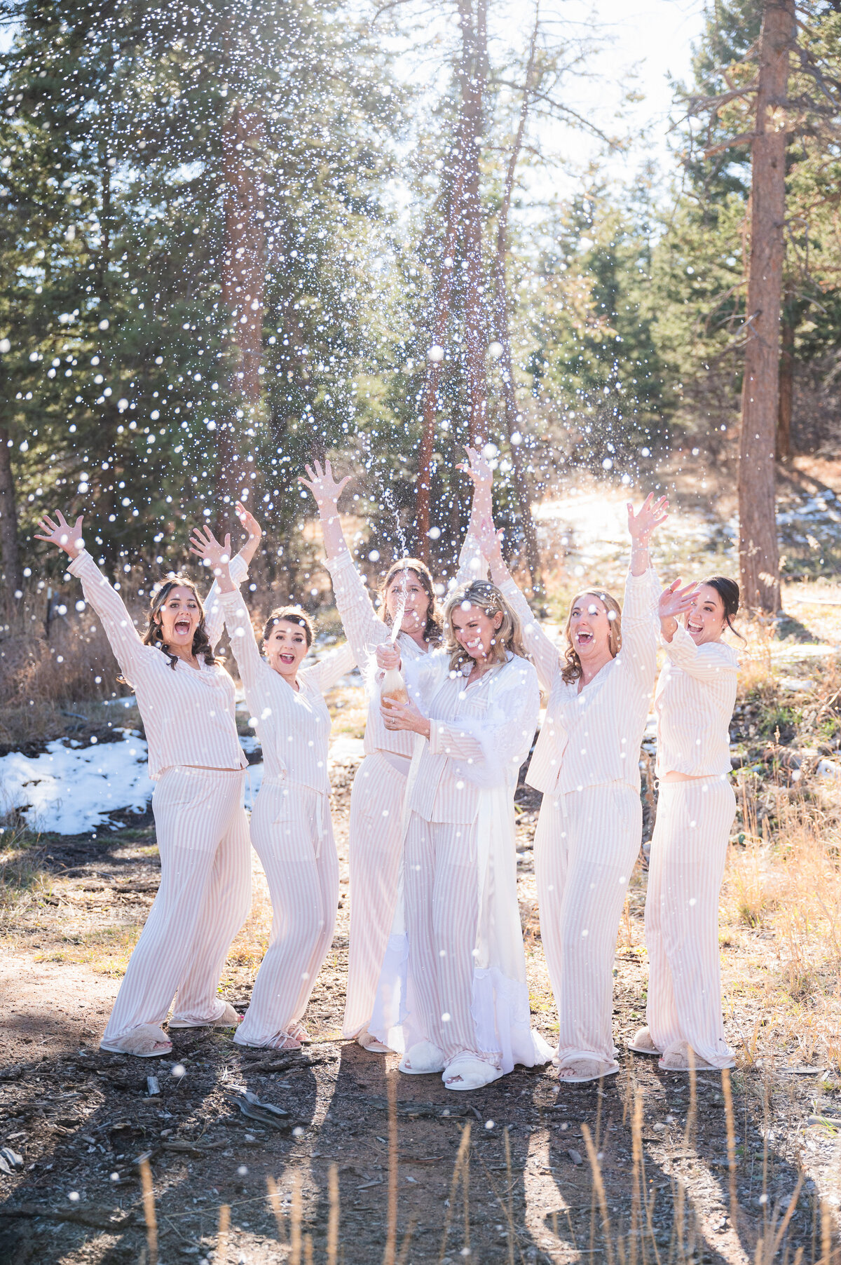 A bride and her bridesmaids pop a bottle of champagne and the bridesmaids all celebrate as bubbles go everywhere.