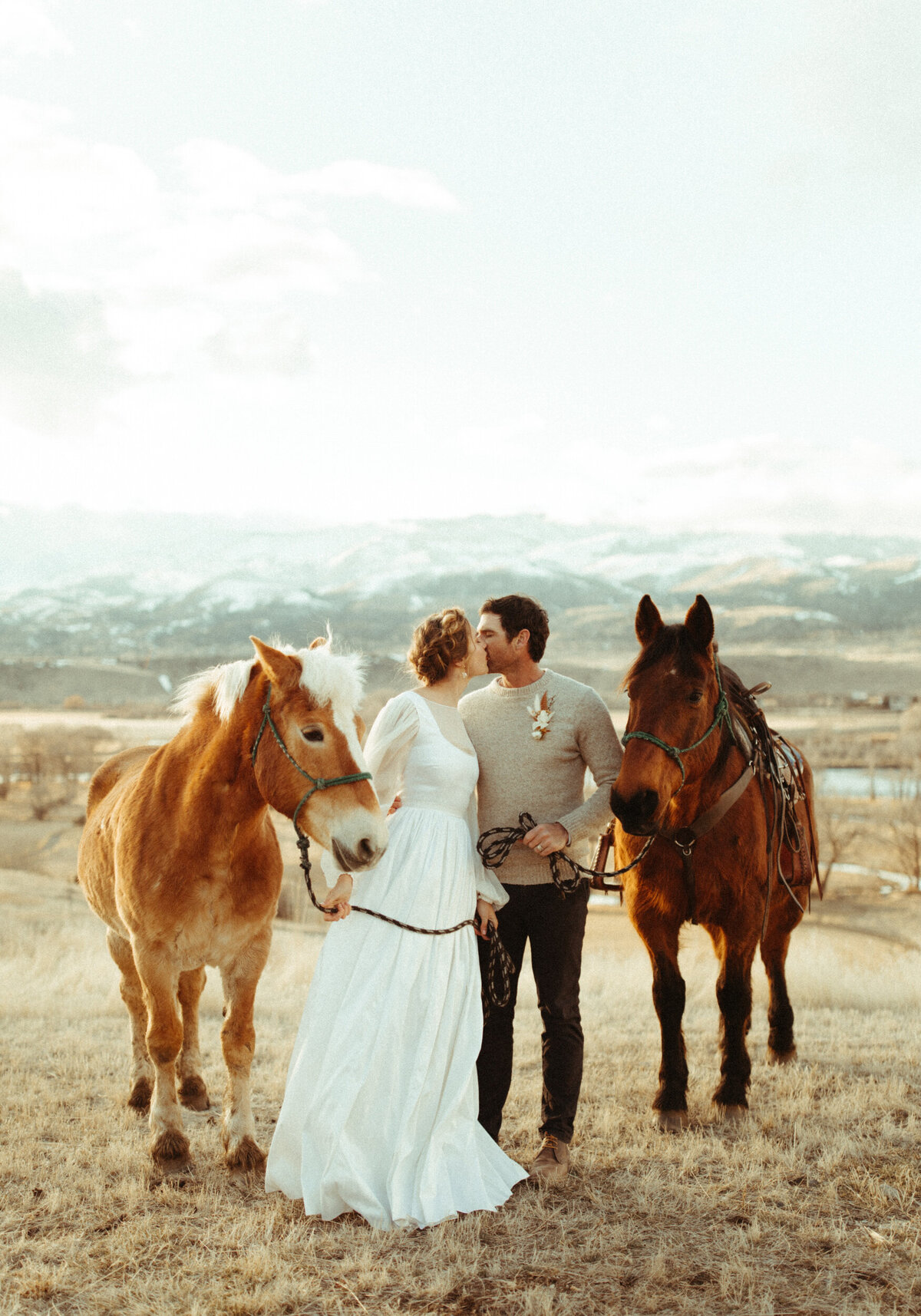 Bride and groom standing between two horses and kissing during a snowy winter mountain elopement