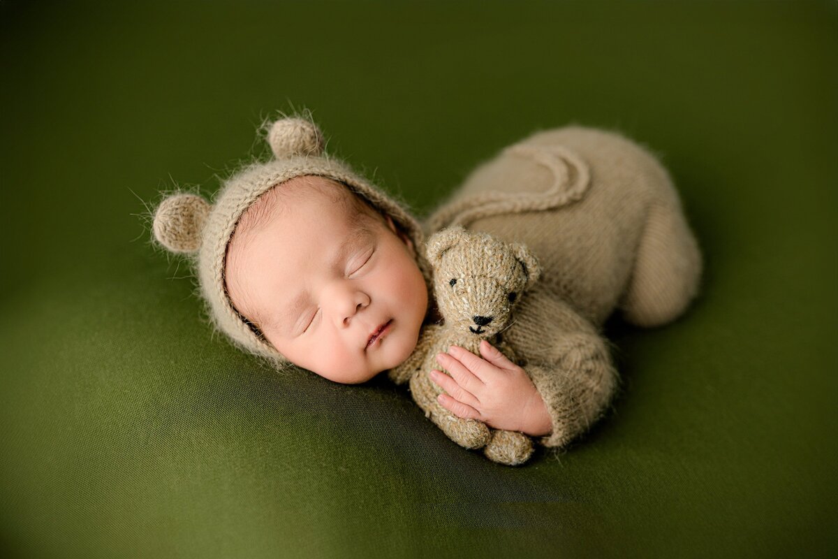 newborn is dressed in a knit outfit and bear bonnet posed in newborn position holding a bear