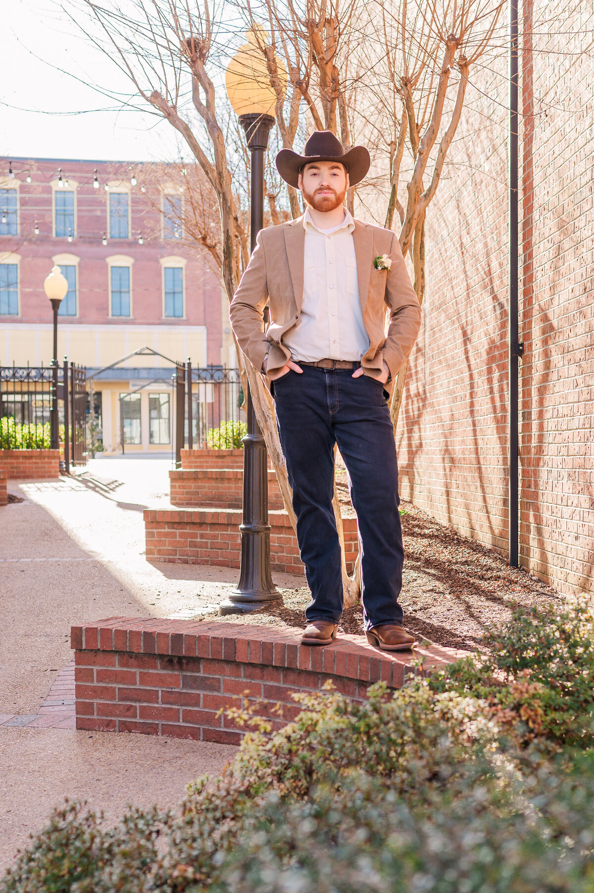 A brunette groom in a  cowboy hat standing on a brick wall in Downtown Oxford enjoying his wedding day with Raleigh wedding photographer, JoLynn Photography
