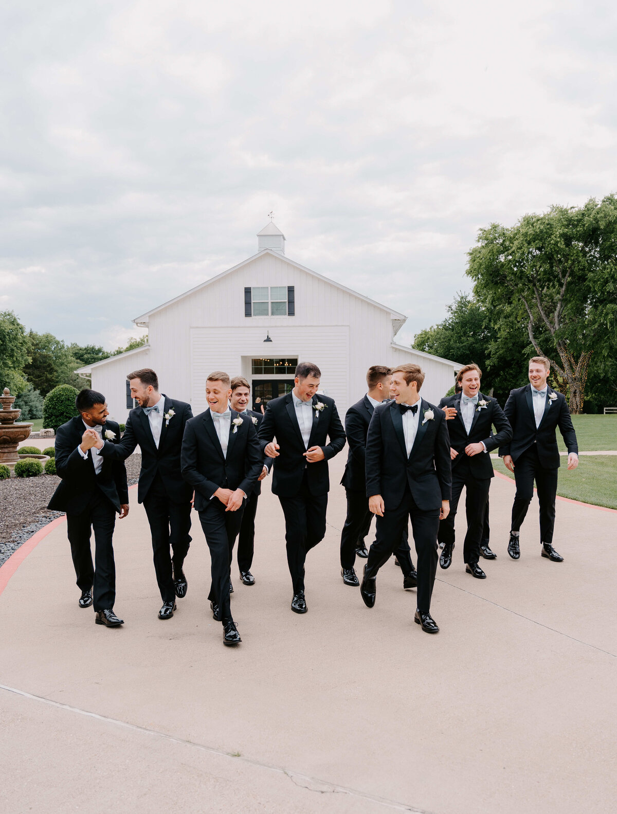 A group of men in black tuxedos walk cheerfully in front of a white barn on a cloudy day. The mood is joyful and celebratory, suggesting a wedding.