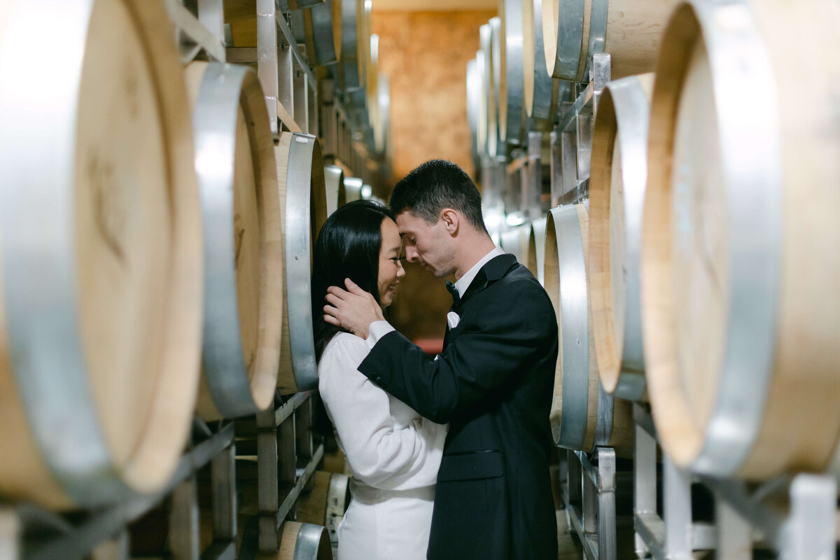 A groom kisses a bride between wine barrels.