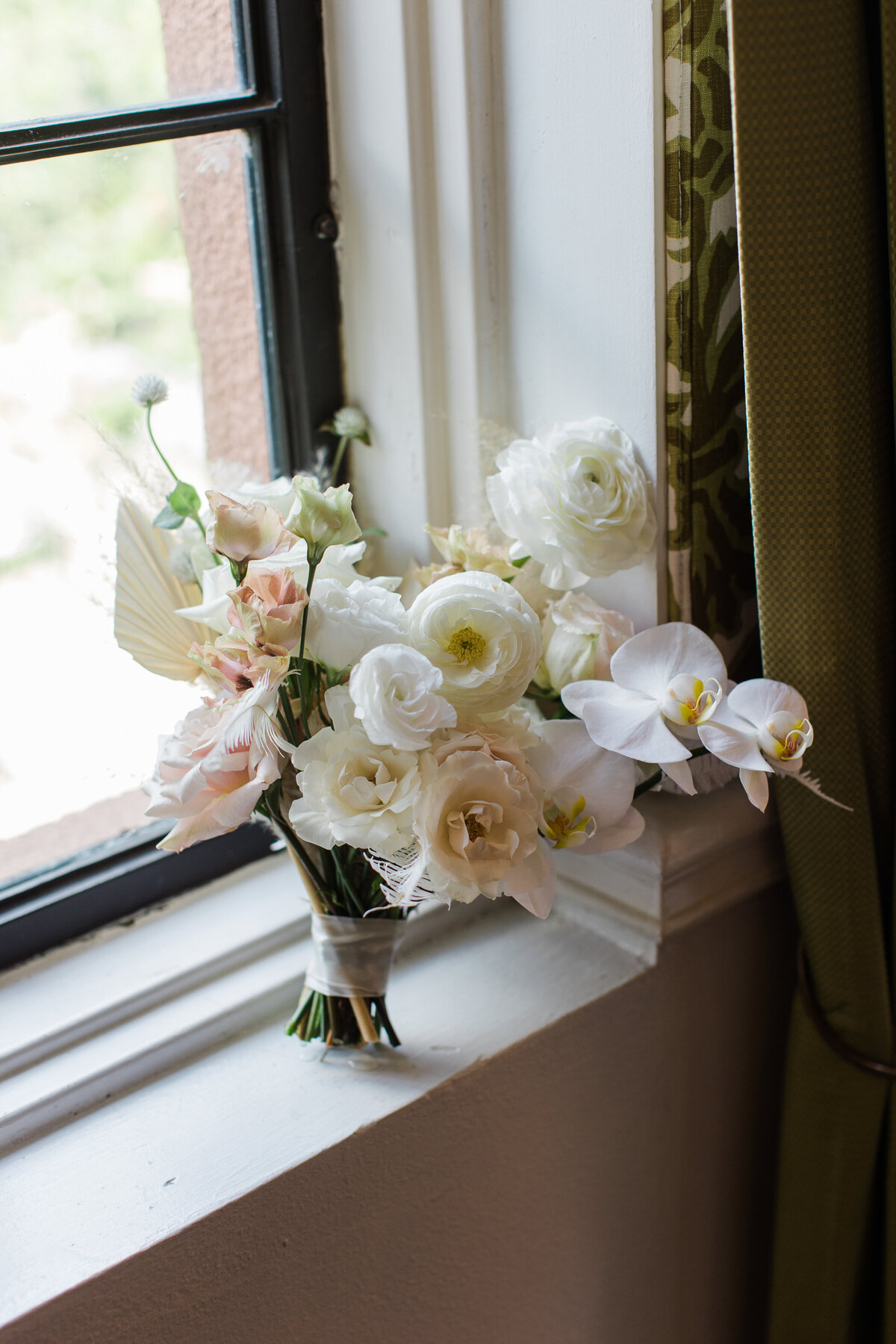 A detail shot of a bridal bouquet for a wedding at the Rosewood Mansion on Turtle Creek in Dallas, Texas. The bouquet is made up of many different types of flowers and contain colors of white, cream, and blush. The bouquet rests on a windowsill next to a intricate set of curtains.