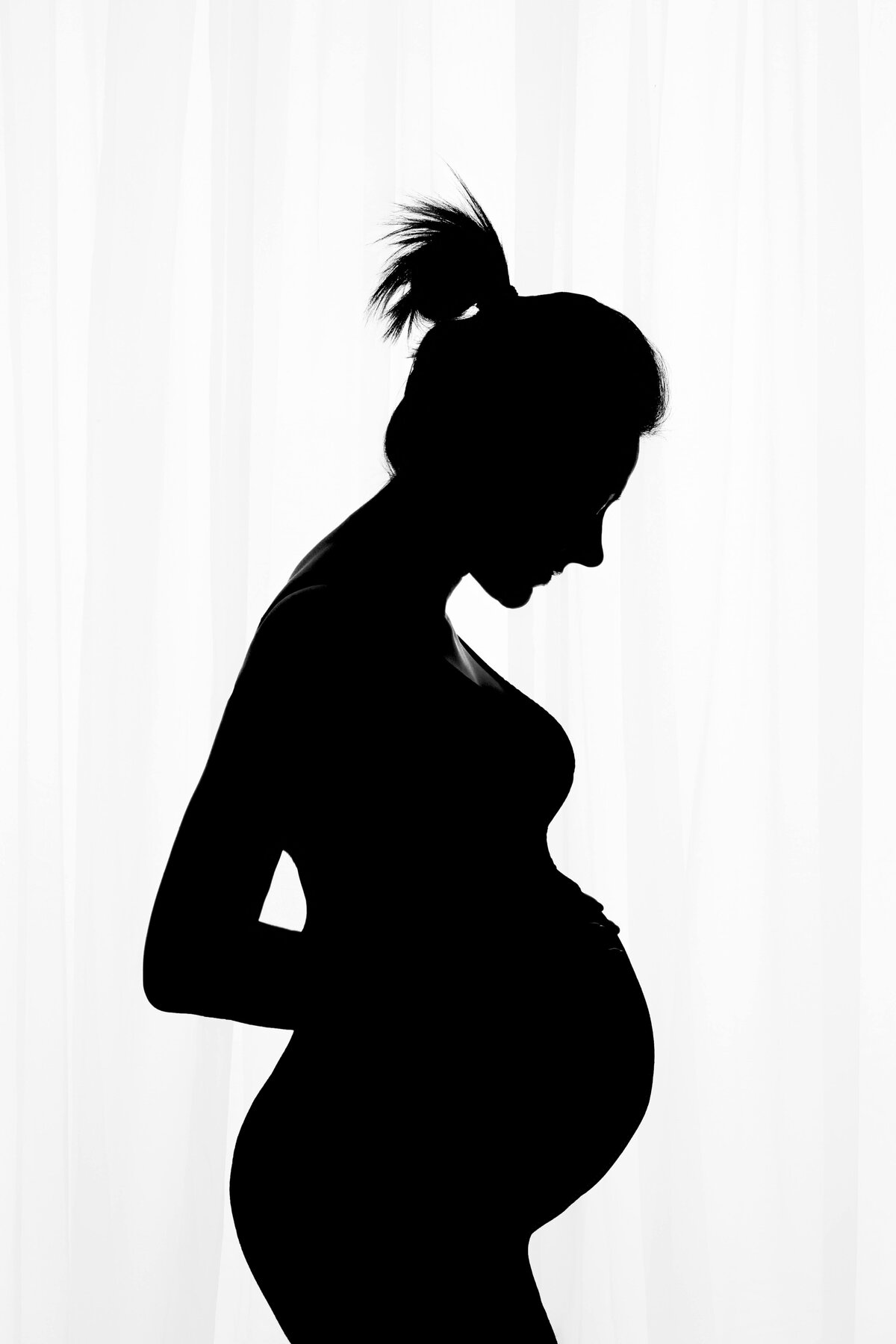 black and white photo of a pregnant woman standing in front of a white back-lit curtain