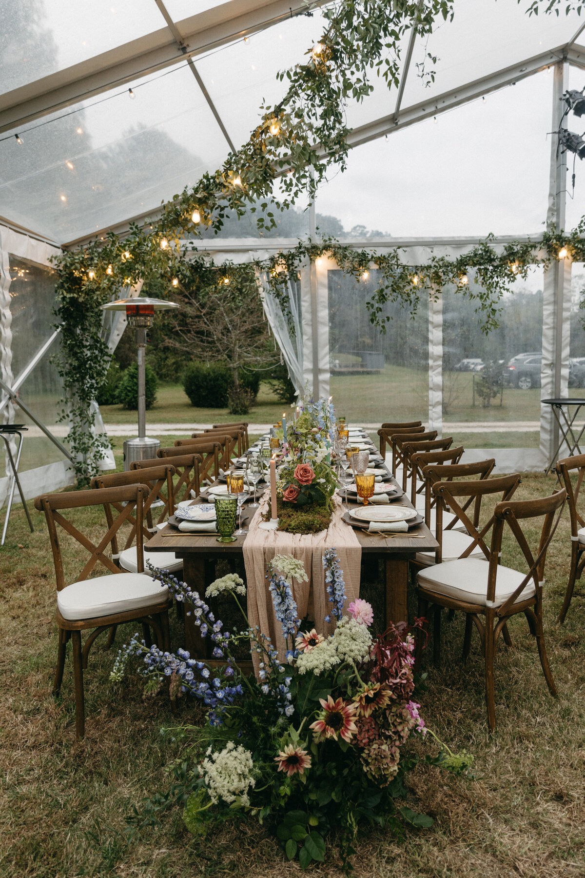 Long family-style reception table lined with whimsical garden-style wedding florals, moss table runner, taper candles, amber glassware, and loose pastel blooms arranged by Fleurish Floral Studio at The Nest in Northwest Arkansas. Clear-top tent draped in greenery and twinkle lights creates an enchanting outdoor dinner setting.