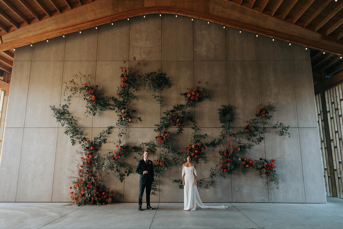 Wedding portrait of groom and bride in front of large industrial modern concrete wall with botanical florals and warm wooden ceiling.