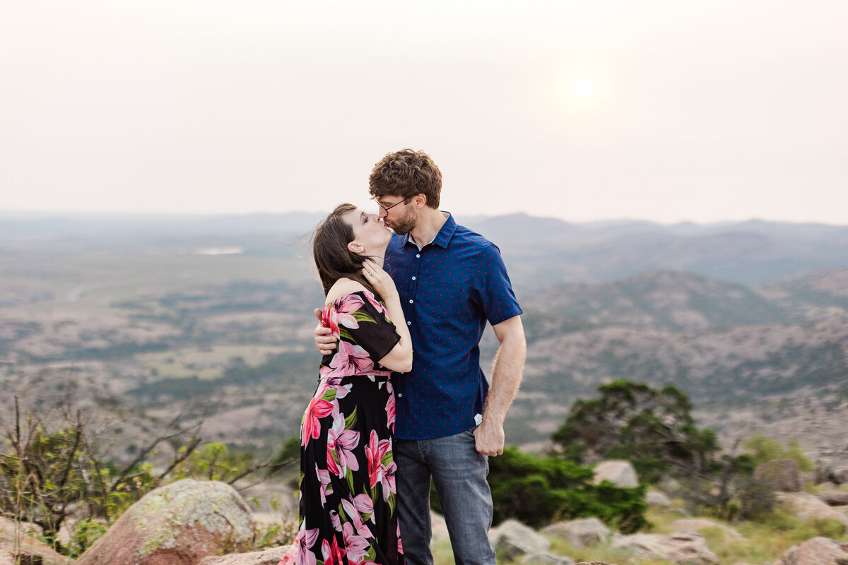 A couple sharing a kiss atop a mountain during their anniversary photoshoot at the Wichita Mountains in Oklahoma. The woman on the left is wearing a black dress covered in large, colorful flowers. The man on the right is wearing a blue dress shirt and jeans. A beautiful landscape can be seen sprawling out behind them.