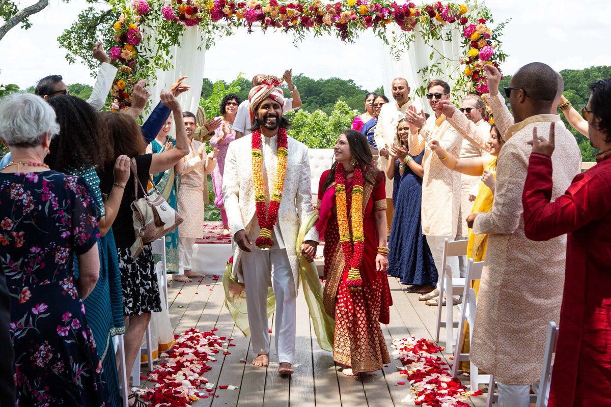 A bride and groom walk under a floral archway, smiling as guests throw flower petals during a traditional Indian wedding ceremony coordinated by a top wedding planner from Des Moines.