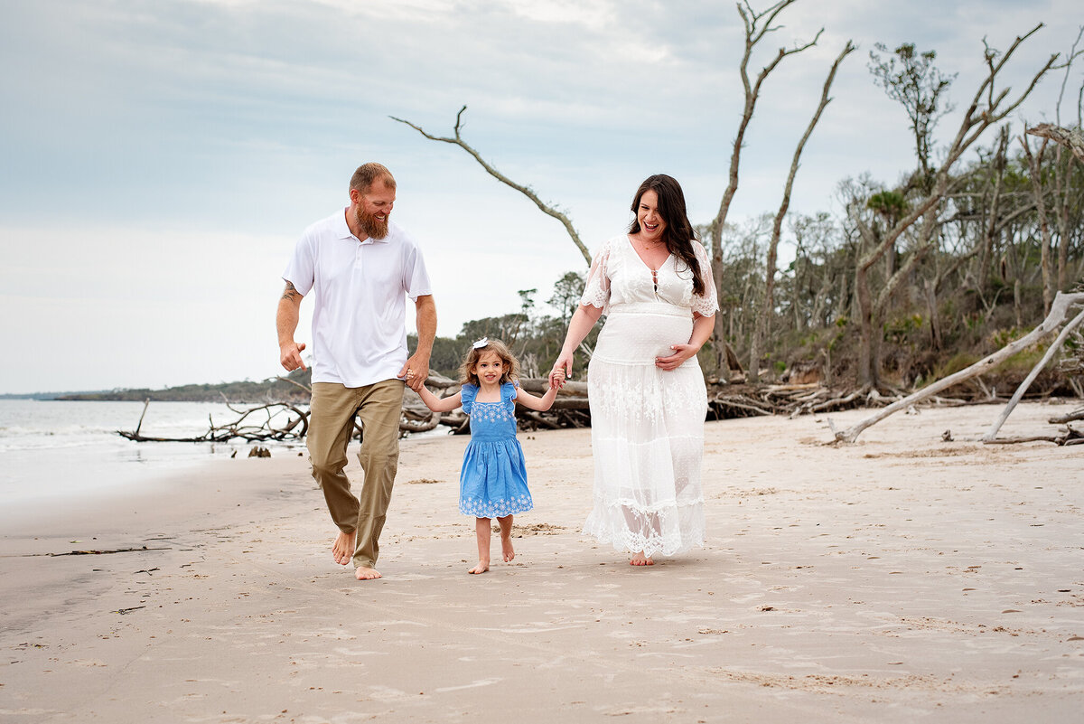 Expectant mom holding hands with daughter and husband on beach in Little Talbot Island, FL.
