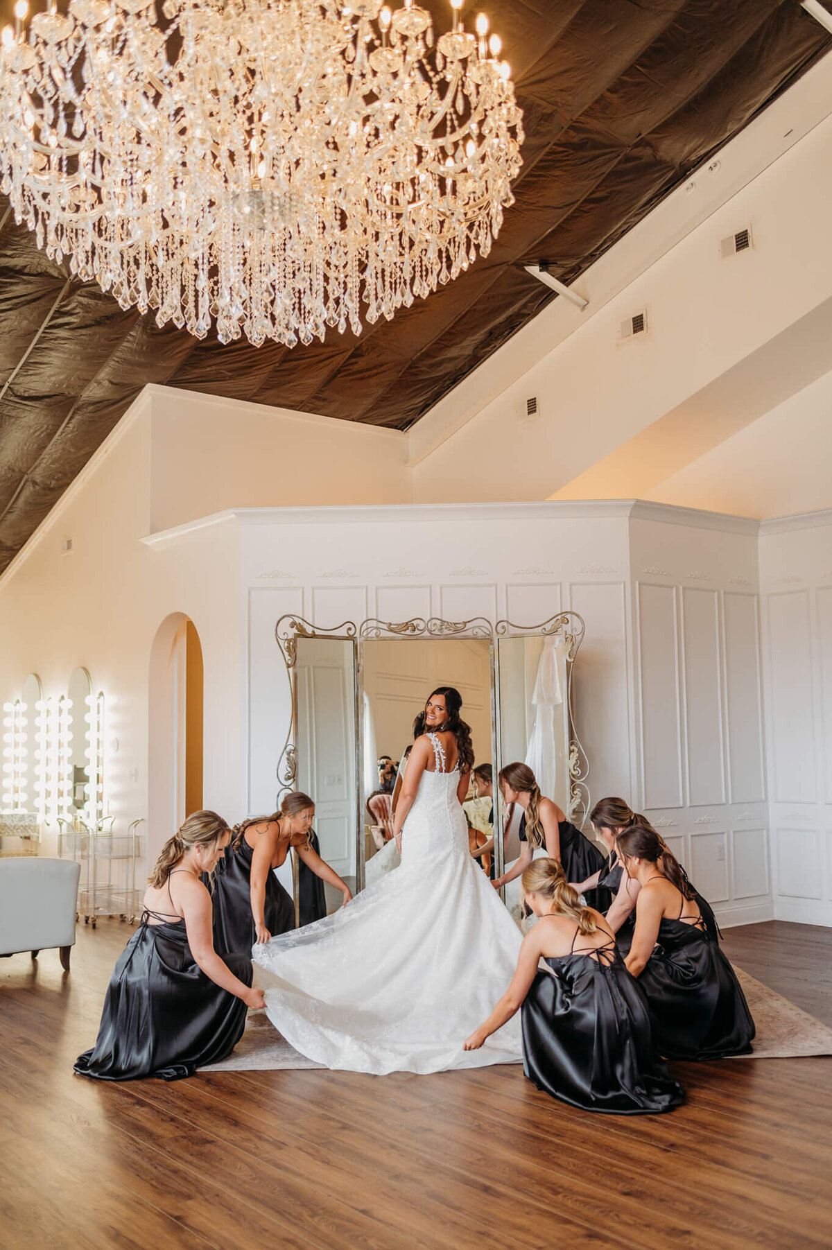 Photo of a bride standing in front of a mirror and under a chandelier as her bridesmaids fix her dress