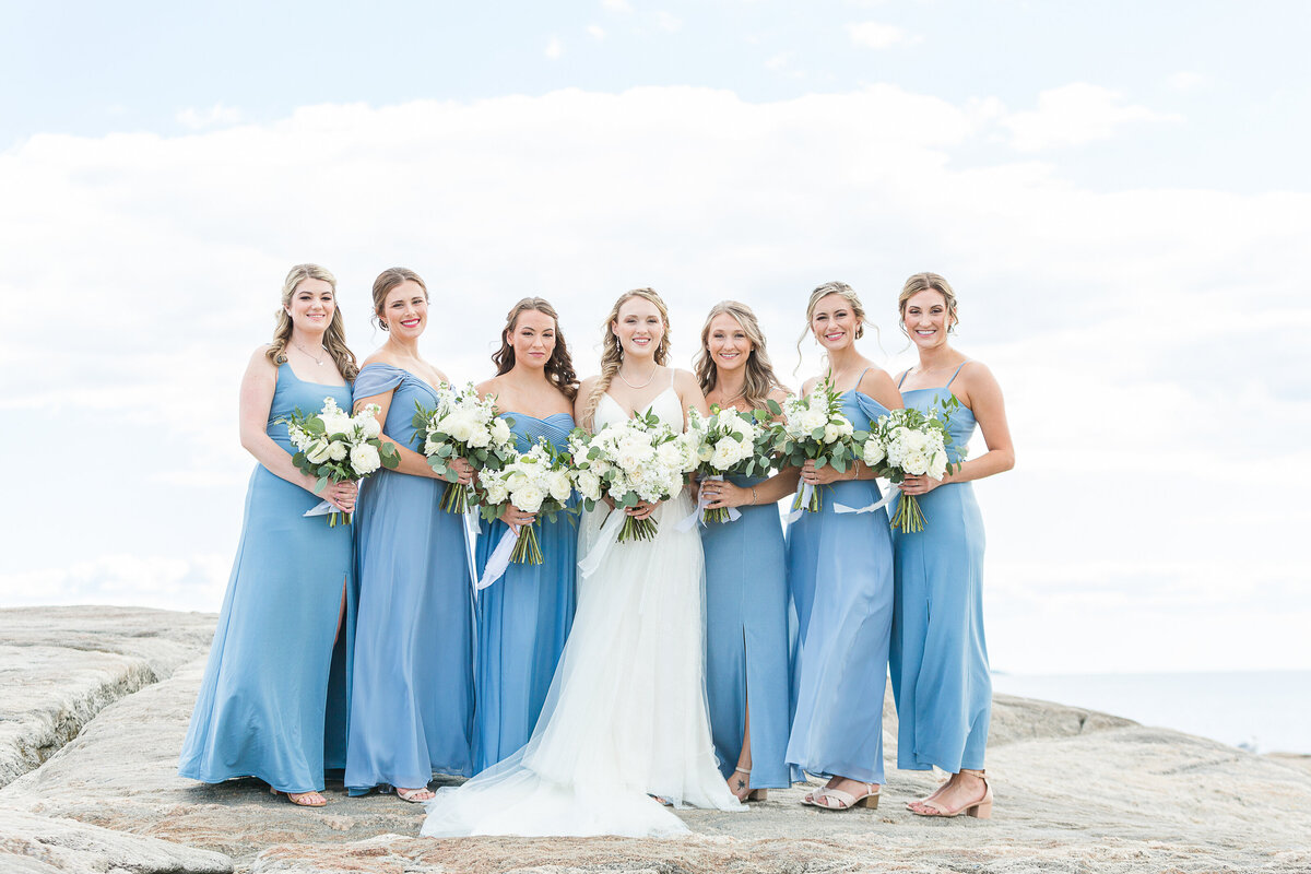 Bride stands on the rocks with her bridesmaids at the Madison Beach Hotel. Bridesmaids in long blue gowns. The ocean is behind them. Captured by best New England wedding photographer Lia Rose Weddings.