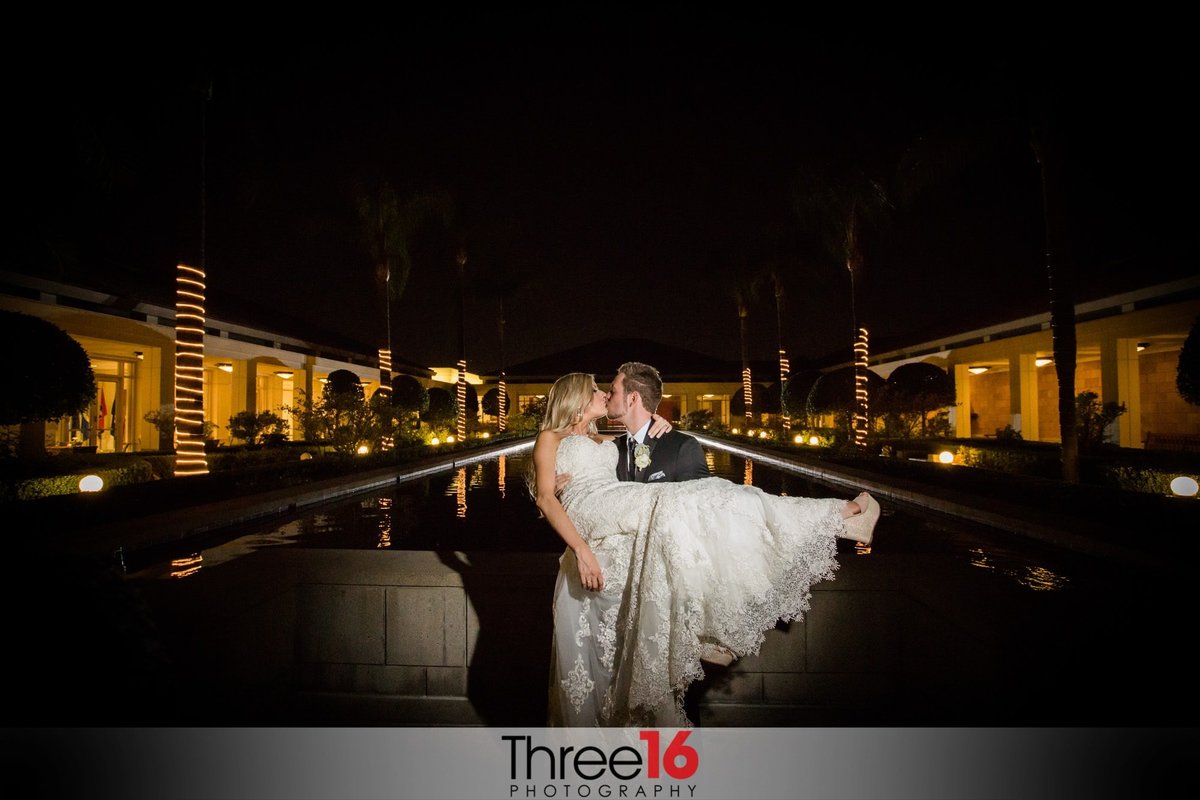 Night shot of the Groom carrying and kissing his Bride next to the reflection pool