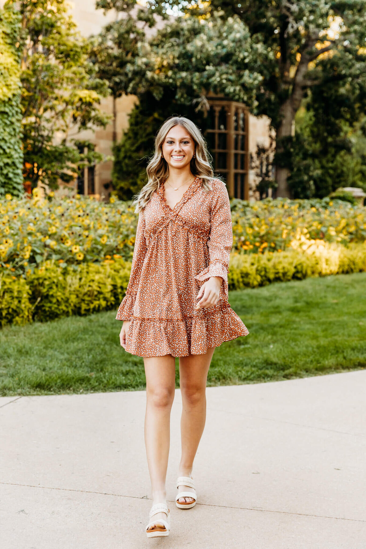 pretty senior girl in a burnt orange boho dress with long sleeves smiling as she walks away from a gorgeous flower bed captured by Ashley Kalbus Photography
