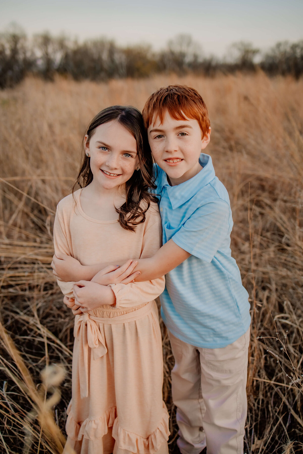 a red haired boy holding onto a brown hared girl both about 8 years old, standing in a field looking at the camera