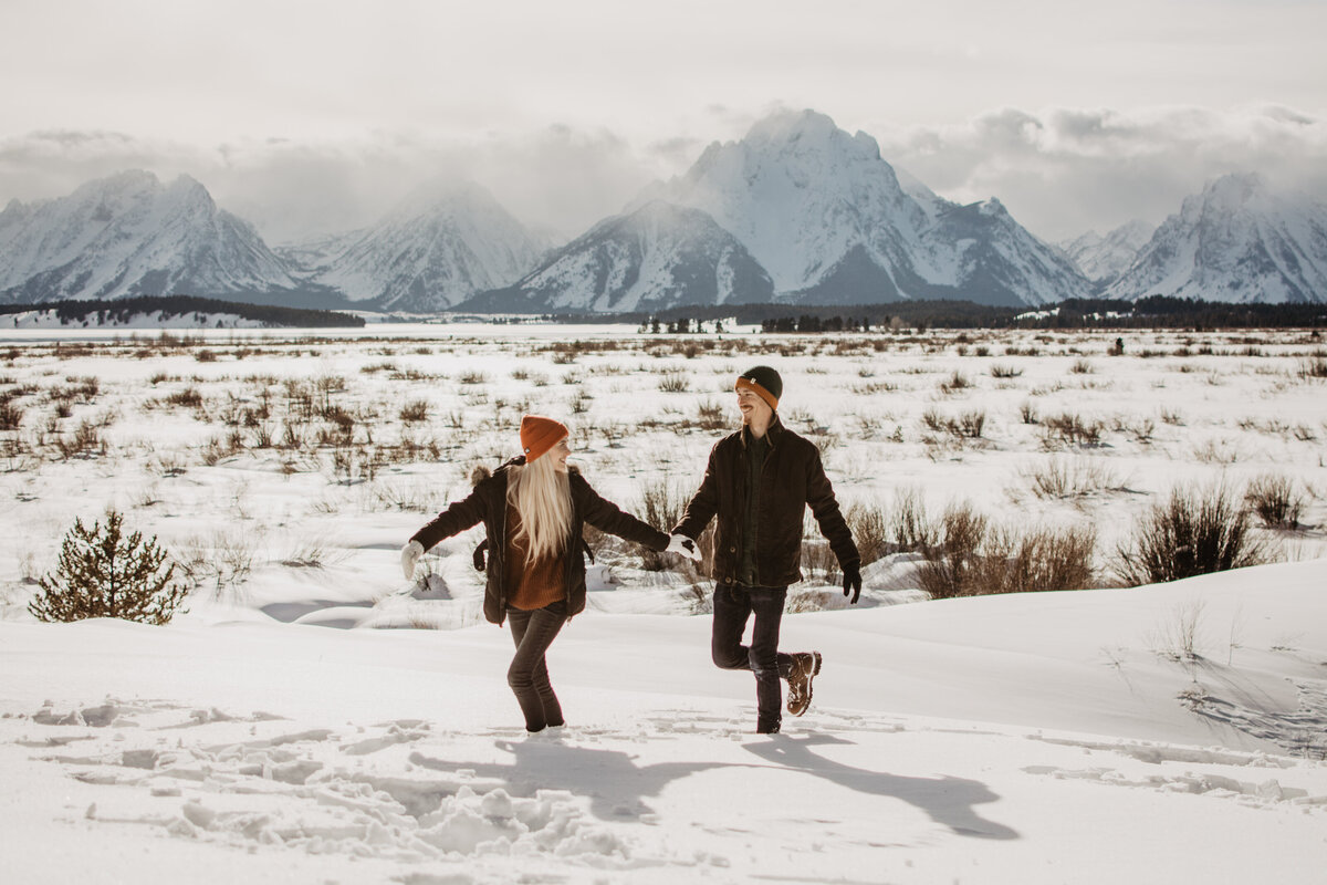 Jackson Hole winter engagement session with man and woman holding hands and running through the snow together in front of the Grand Tetons captured by jackson hole photographers