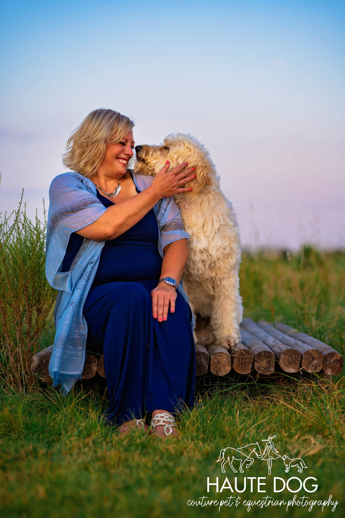 A woman and a Doodle dog with curly fur touching noses affectionately, with the woman wearing a smile on her face..