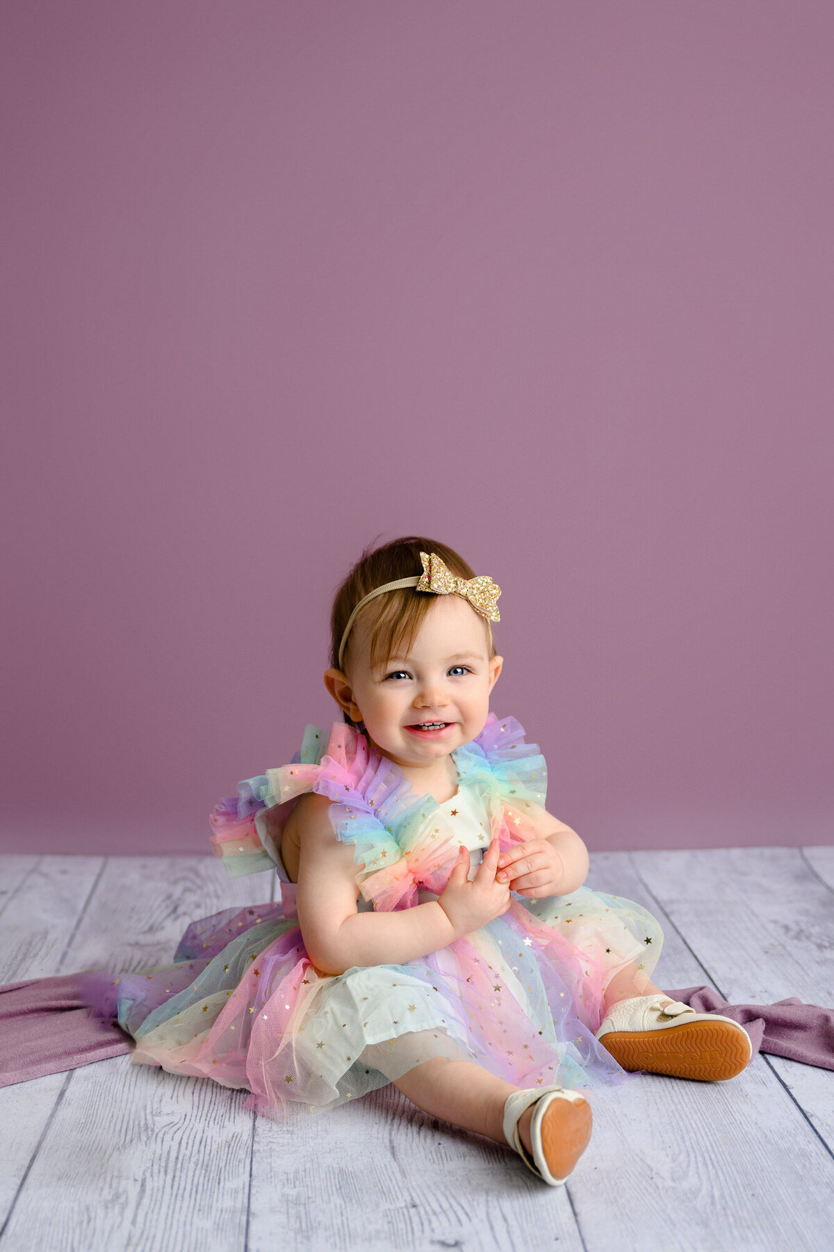 Little girl is dressed in a rainbow dress sitting on the ground during her 1-year-old Milestone session with a purple background