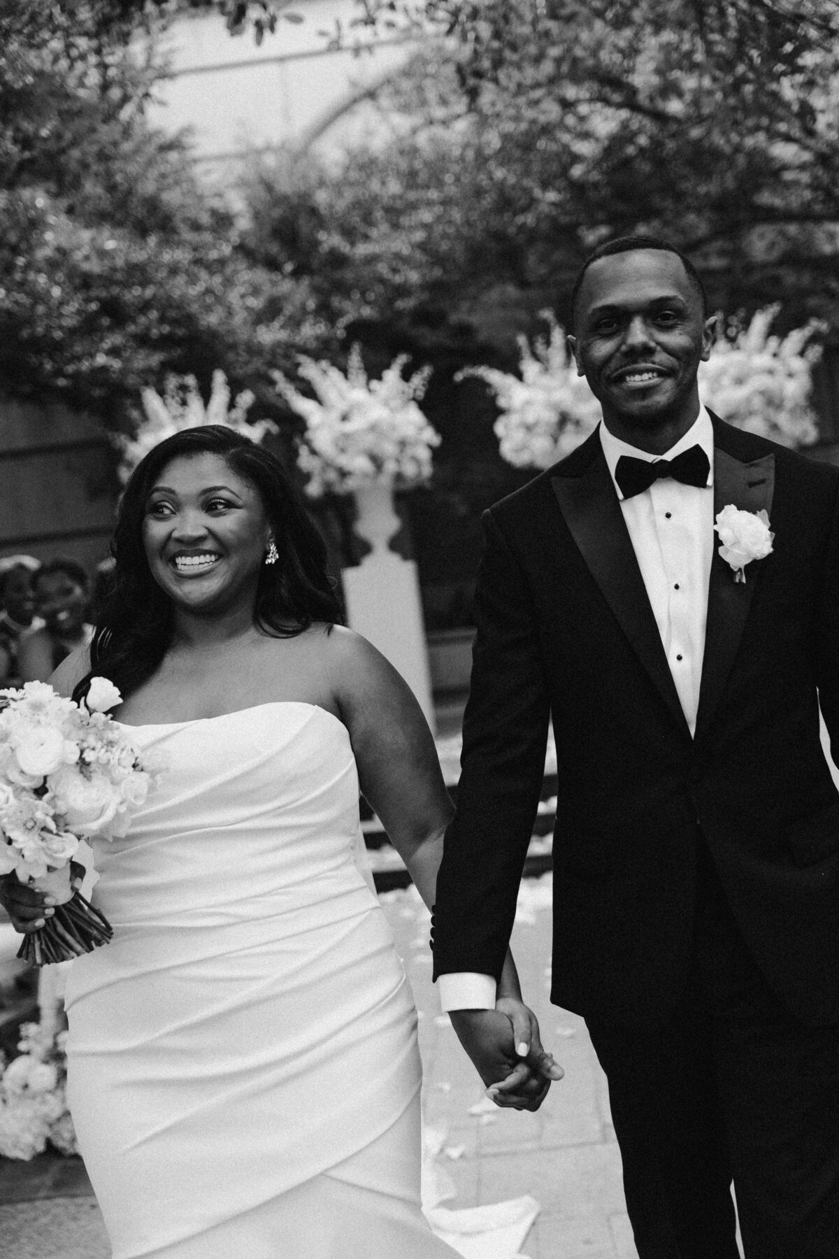 A joyful couple walks hand in hand at their wedding. The bride in a white gown holds a bouquet, and the groom in a black suit smiles warmly.