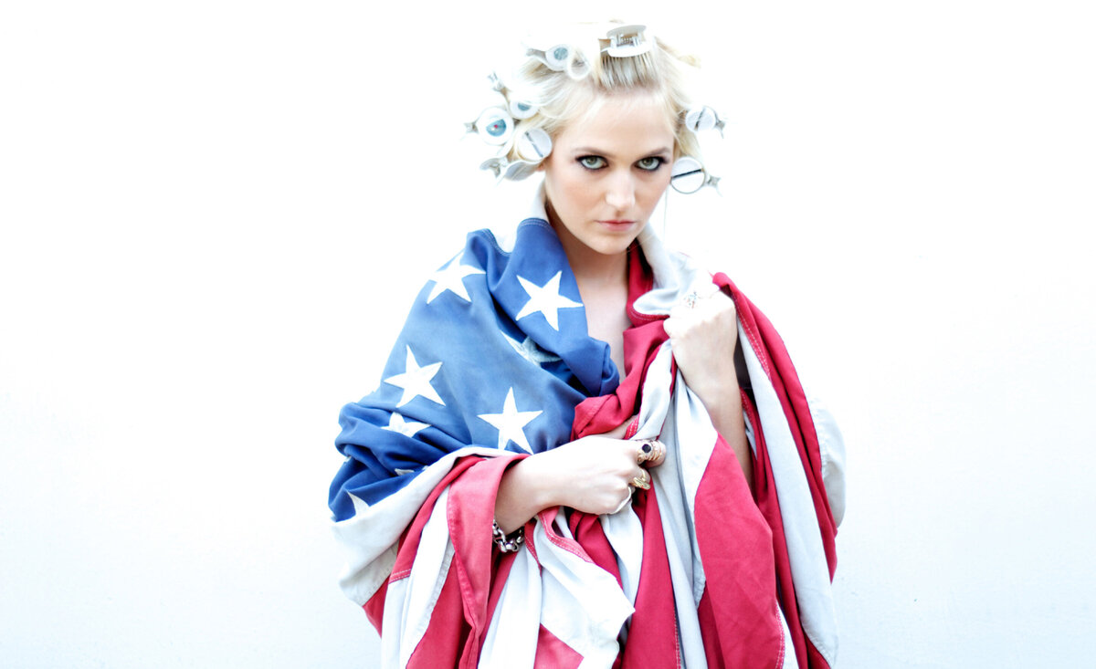 Women music portrait Nikki Paige wrapped in large american flag against white wall
