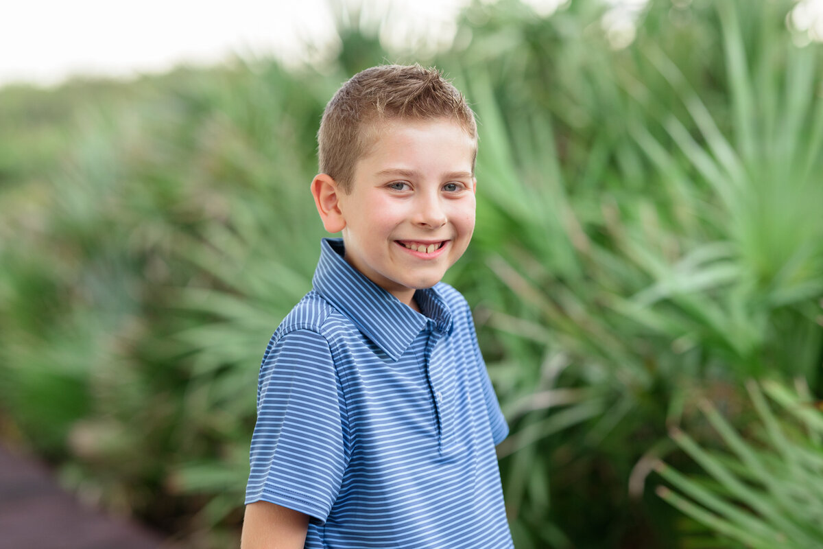 A young boy smiling with green plants behind, on a boardwalk at Rosemary Beach.