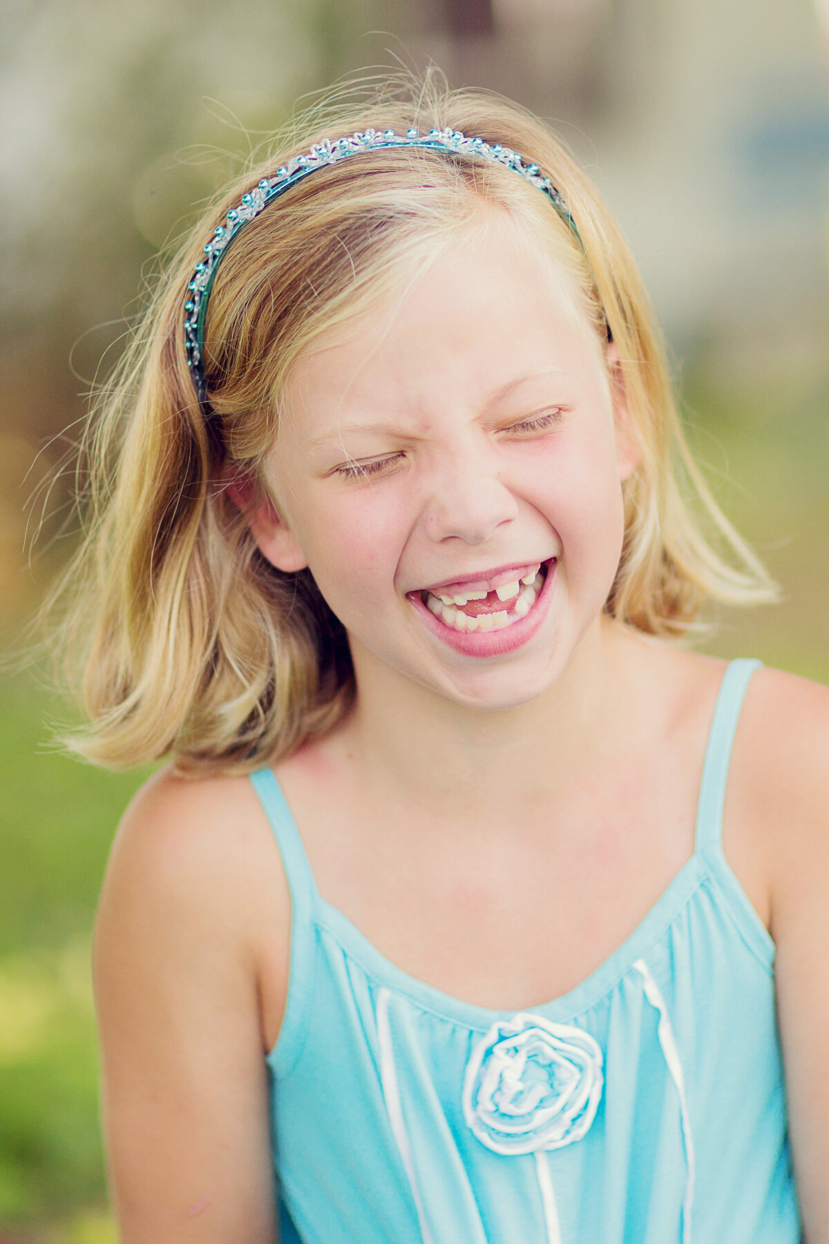 Girl giggles with eyes closed, showing off her crooked teeth coming in. She's wearing a light blue top and a matching headband.