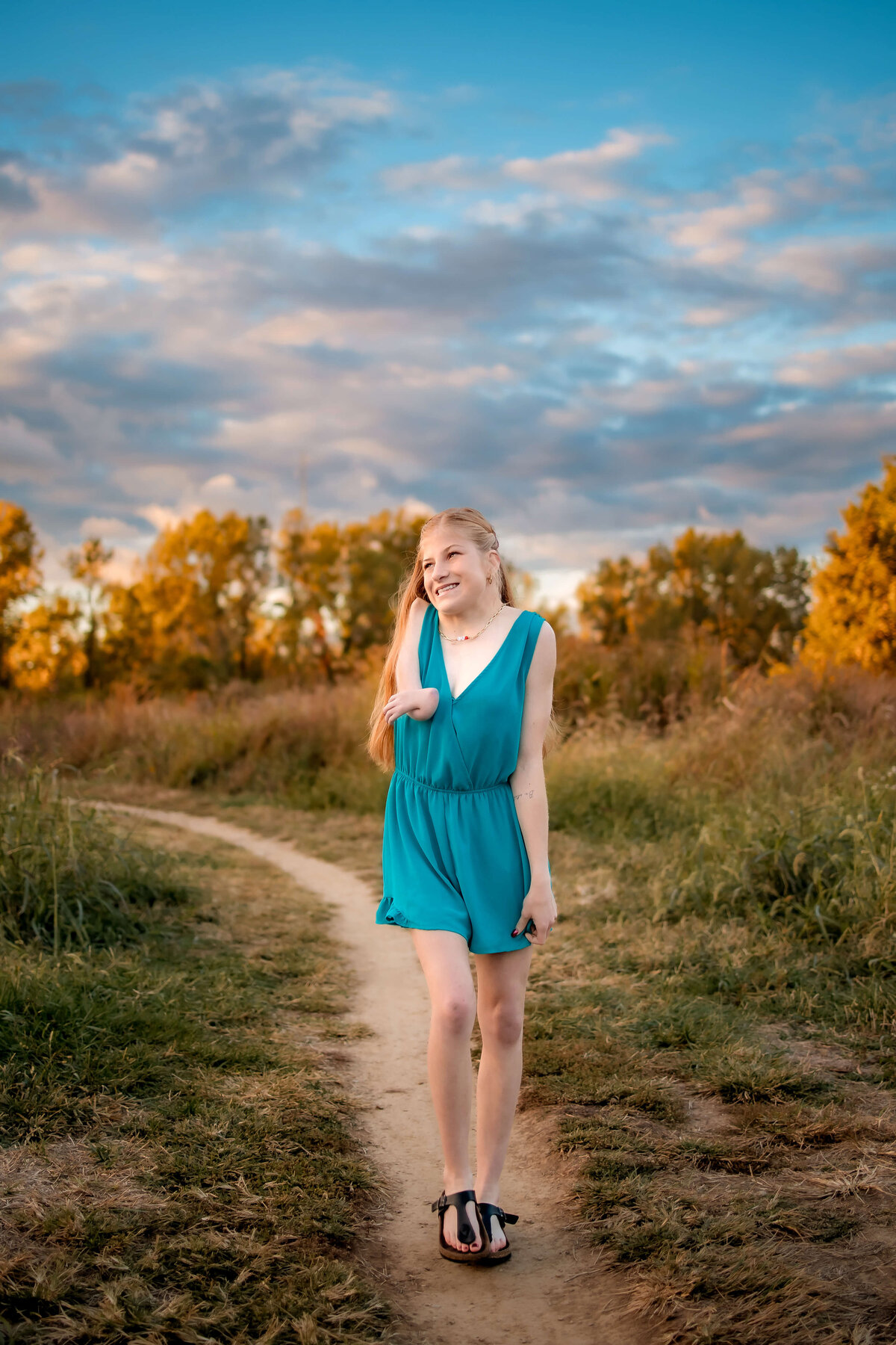 A gorgeous young lady is walking on a dirt path in a park on a fall day with a beautiful and colorful sky filled with clouds for her senior portraits.
