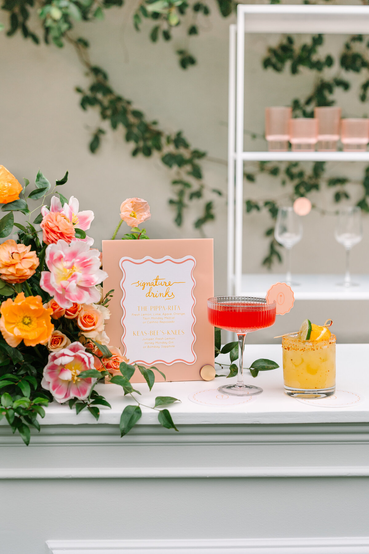 wedding bar with orange and pink florals, two colorful cocktails, and signature drink menu.
