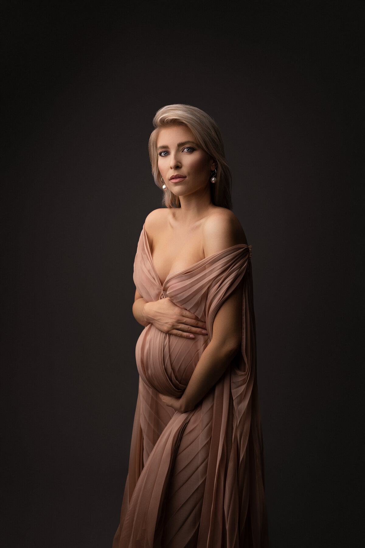 Woman poses for fine art maternity photos with top Philadelphia Main Line maternity photographer Katie Marshall. The woman is standing angled to the camera in a long, off-the-shoulder dusty rose gown with floor-length caped sleeves. One and is atop of her baby bump, the other is under. She is looking over her shoulder toward the camera with an intent gaze.