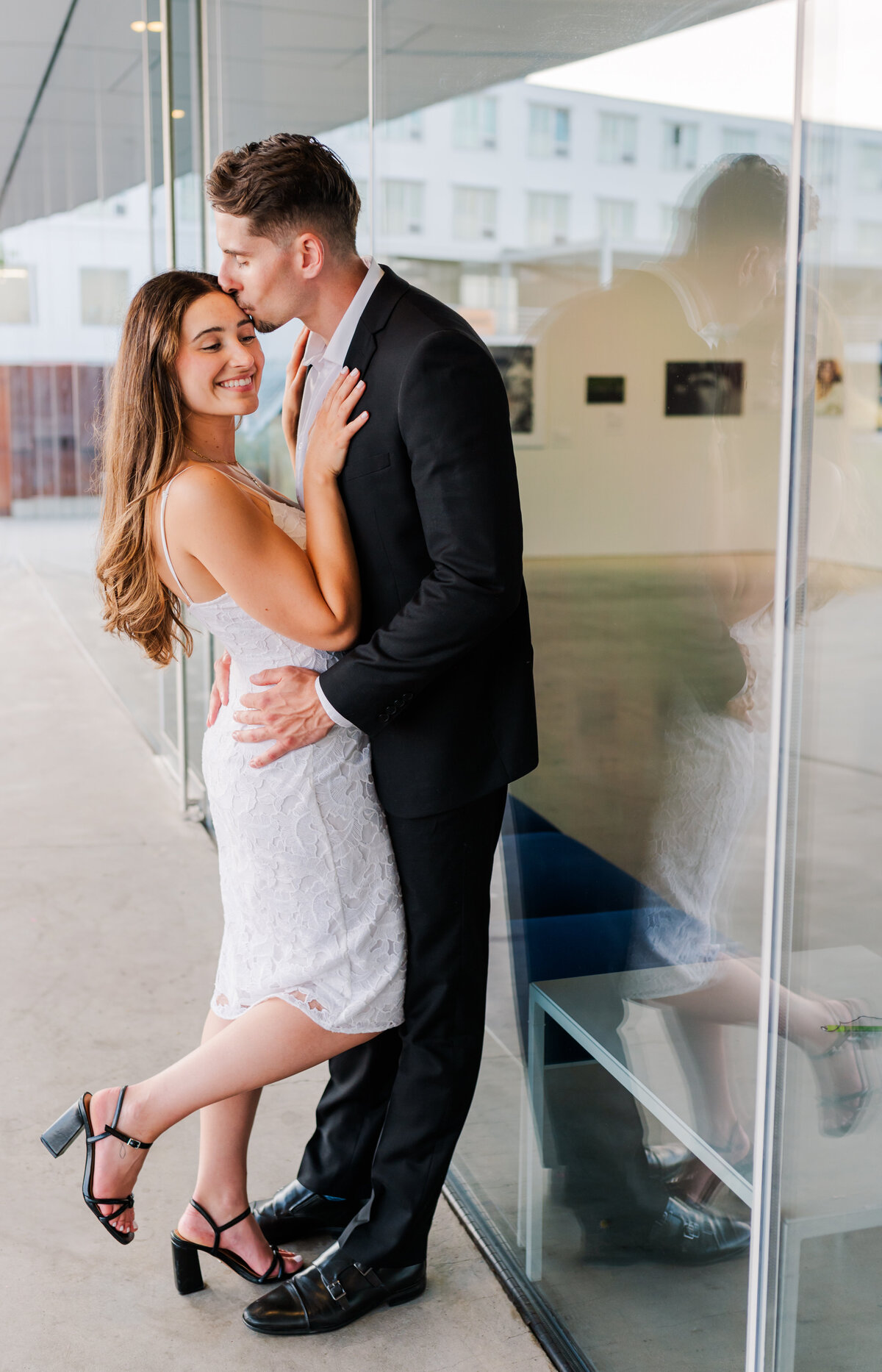 Guy leans against a glass window girl is close to the guy one leg popped up and the guy kisses the girl on the forehead, girl looking down. Both dressed in a black & white attire.