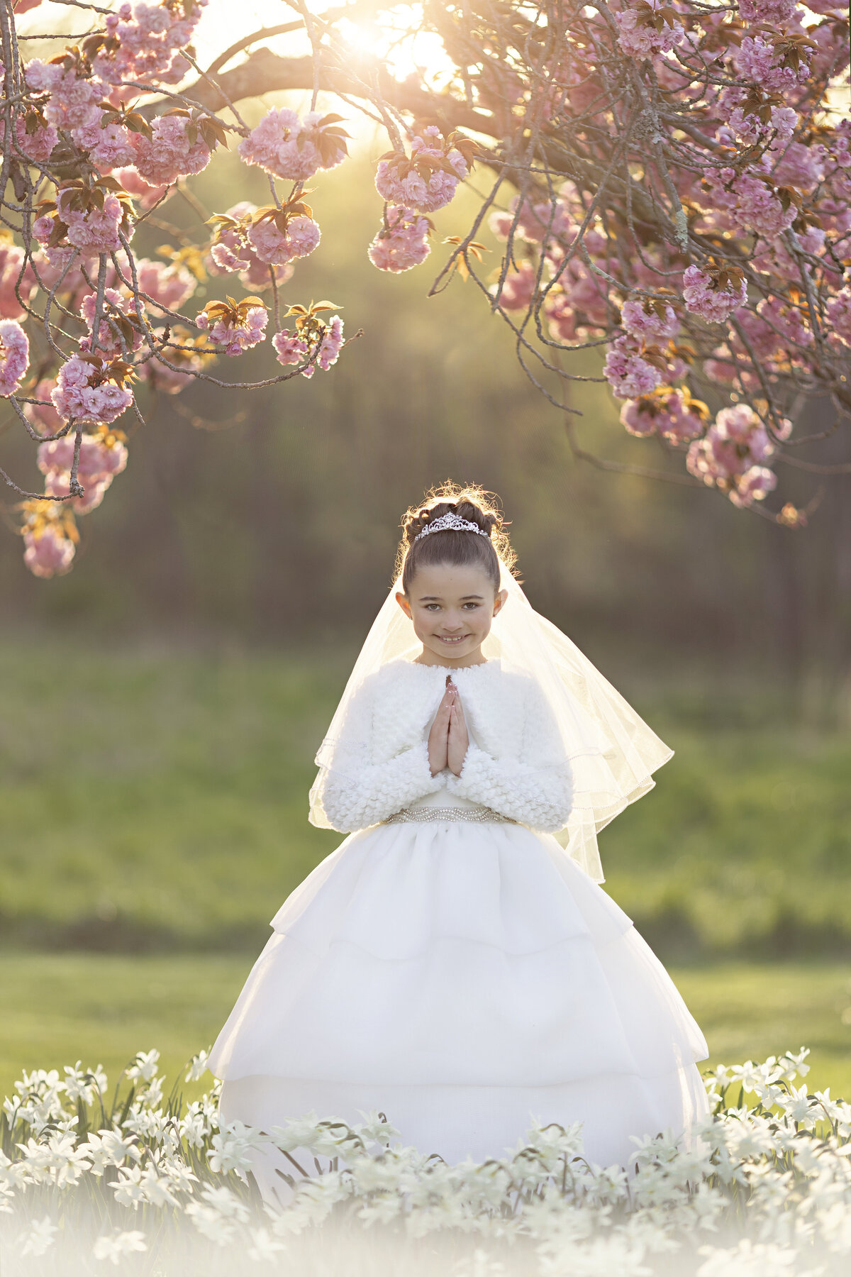 A young girl prays in her communion dress and tiara among white wildflowers
