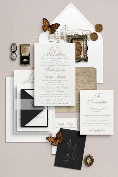 A romantic wedding invitation suite displayed against a soft nude/taupe background. The main invitation, printed on ivory white paper, showcases a delicate floral crest with a custom monogram and romantic script typeface. The printed envelope liner features a custom venue sketch, complemented by black RSVP envelopes with metallic gold ink accents. The suite is adorned with custom wax seals and vintage ring boxes.
