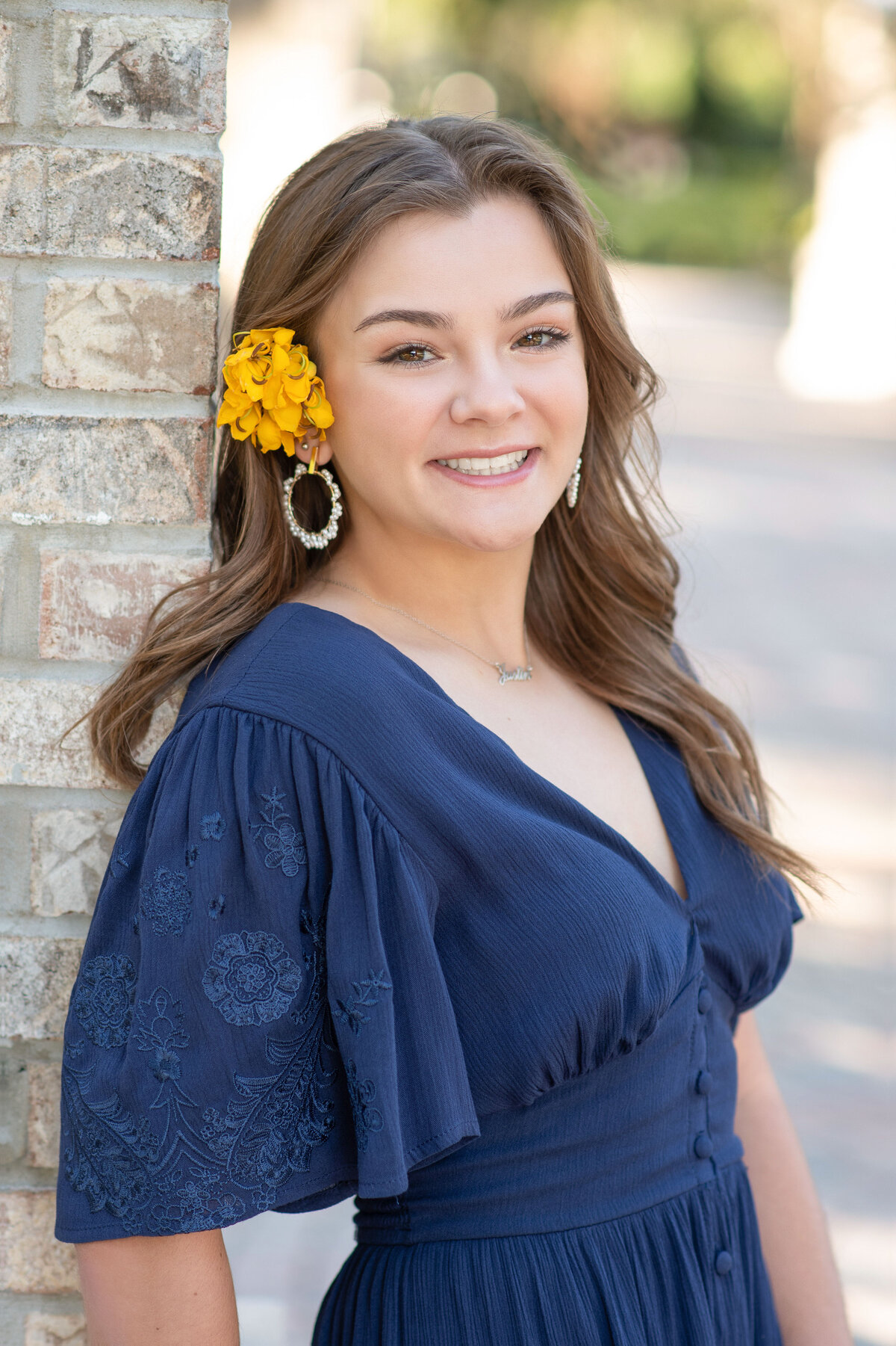 High school senior girl with yellow flowers in hair leans against a brick pillar.