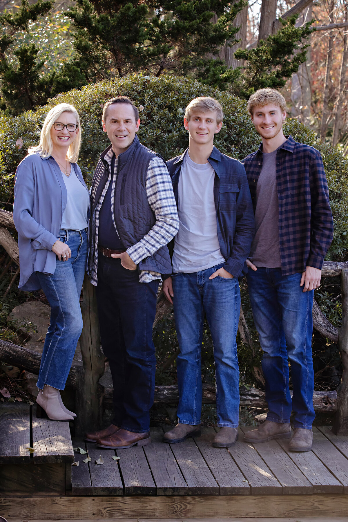 Picturesque family of four in casual fall attire posed by rustic wooden railing with lush foliage backdrop at UNCC Botanical Garden
