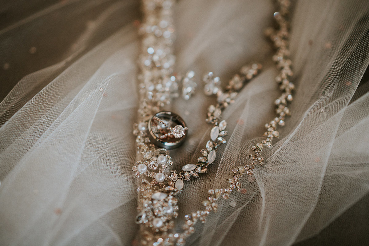 Round diamond engagement ring inside wedding rings on top of pearl bridal jewelry on top of rhinestone wedding veil