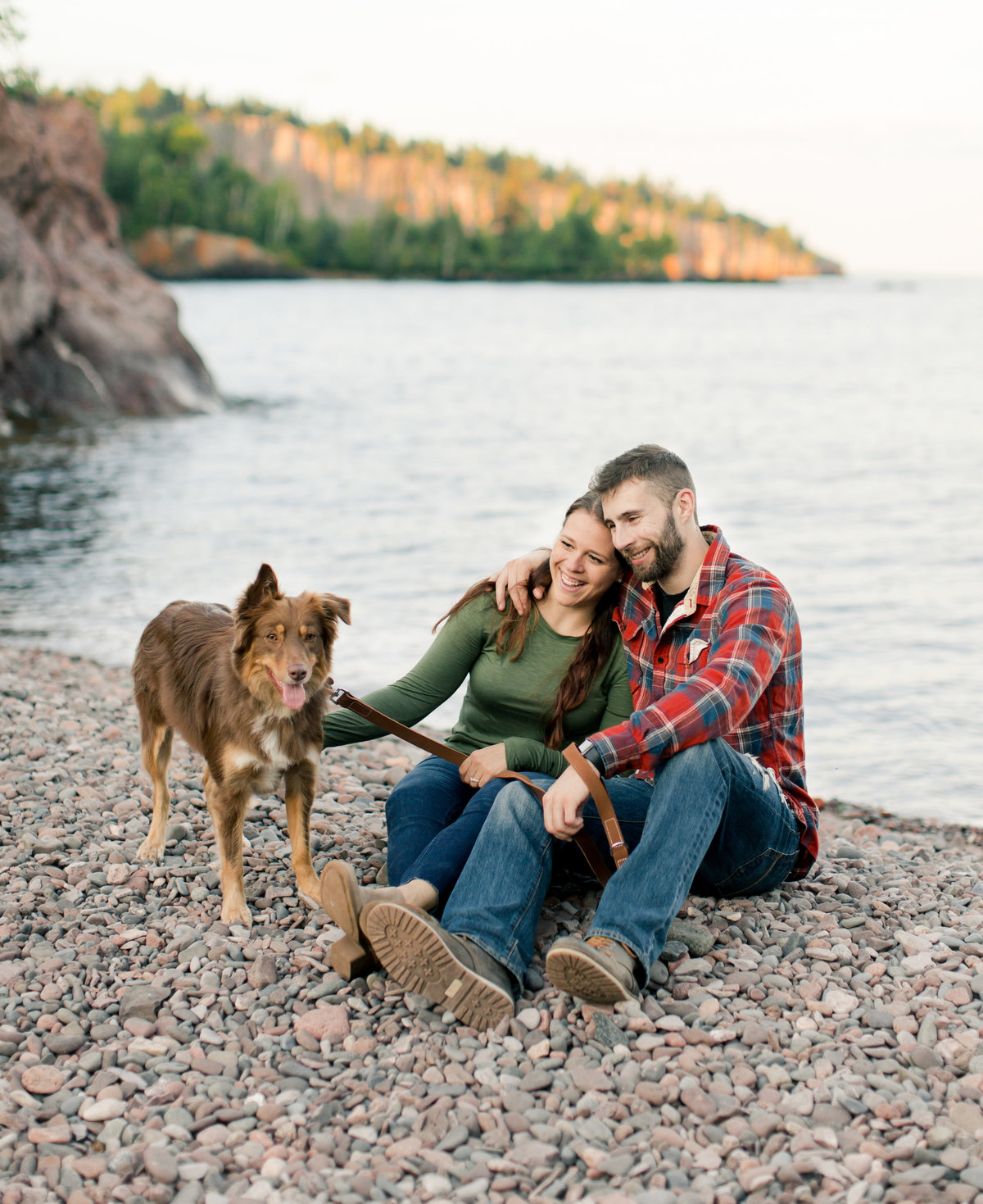 couple sits on a pebble beach with dog along shores of lake superior