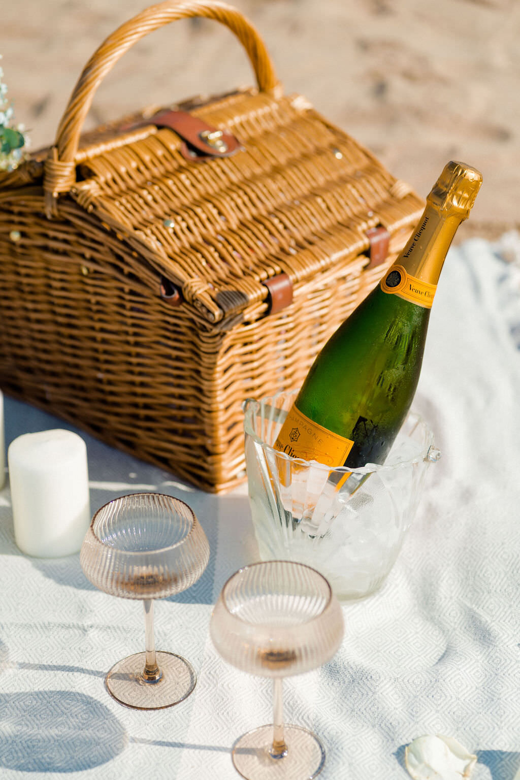 champagne glasses, a champagne bottle on ice, candles, and a picnic basket laid out on a blanket