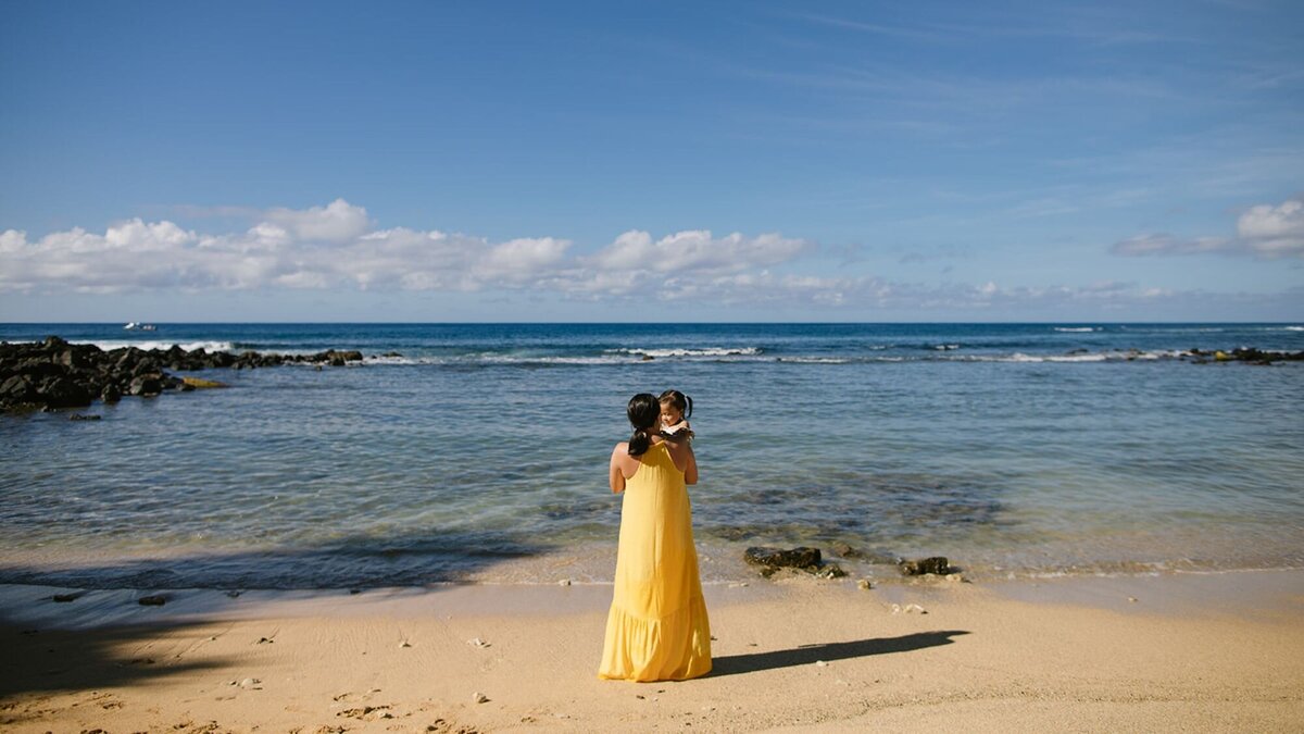 A woman in a yellow dress holds her baby on the beach of Kauai.