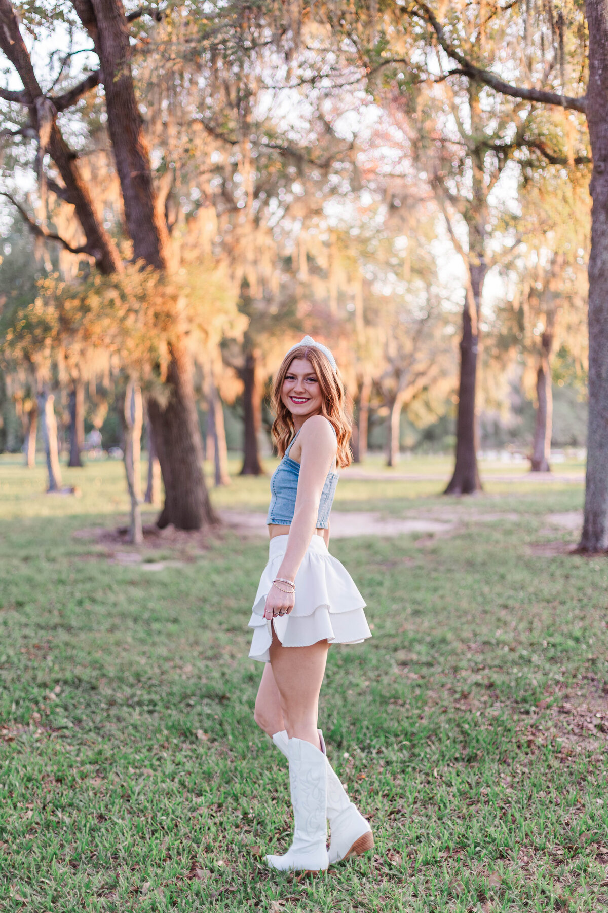 A cowgirl from santa fe high school stands in a field with mossy trees while wearing a denim vest, white leather skirt, and white cowboy boots