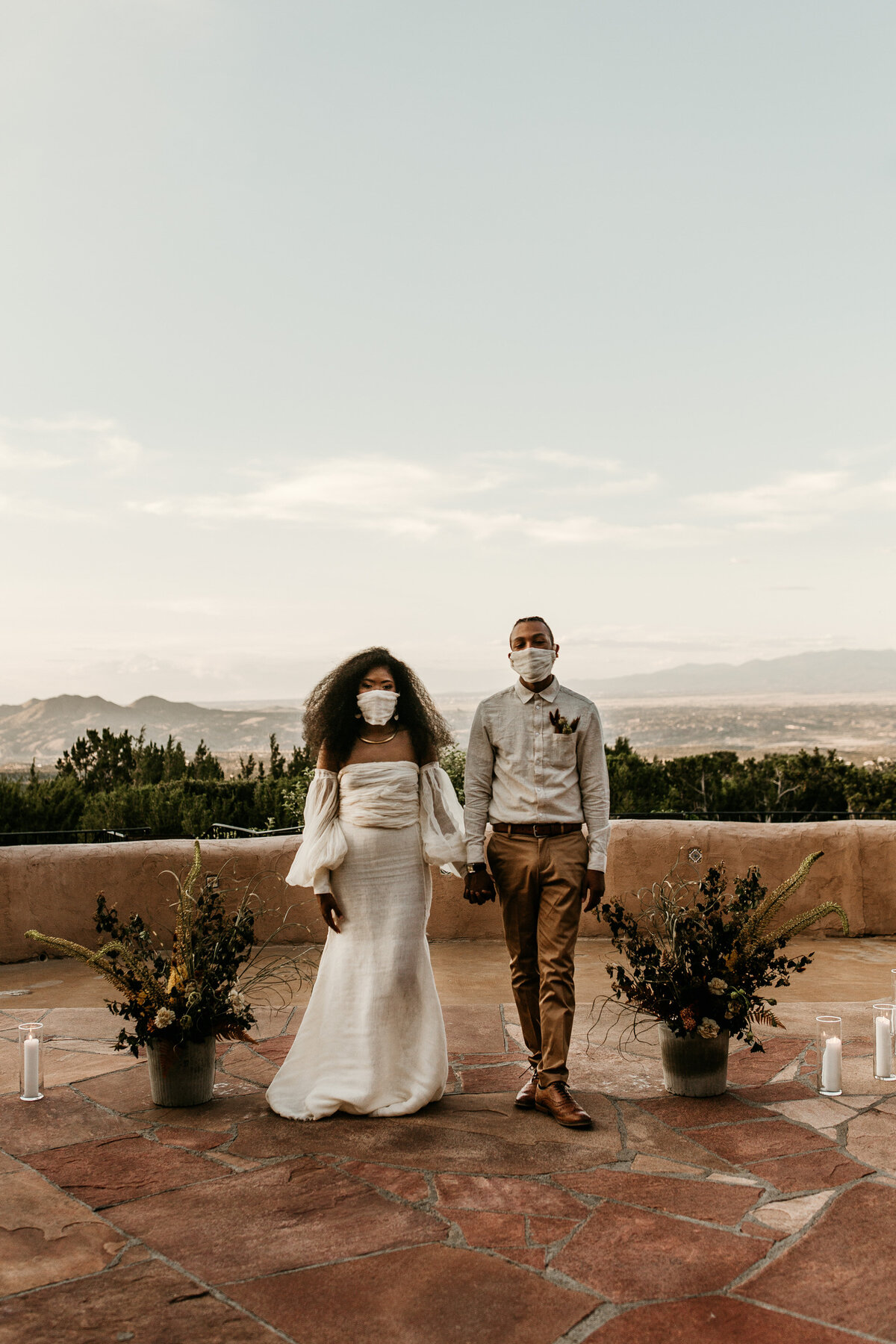 bride and groom holding hands wearing masks at Santa Fe venue courtyard with mountains behind them