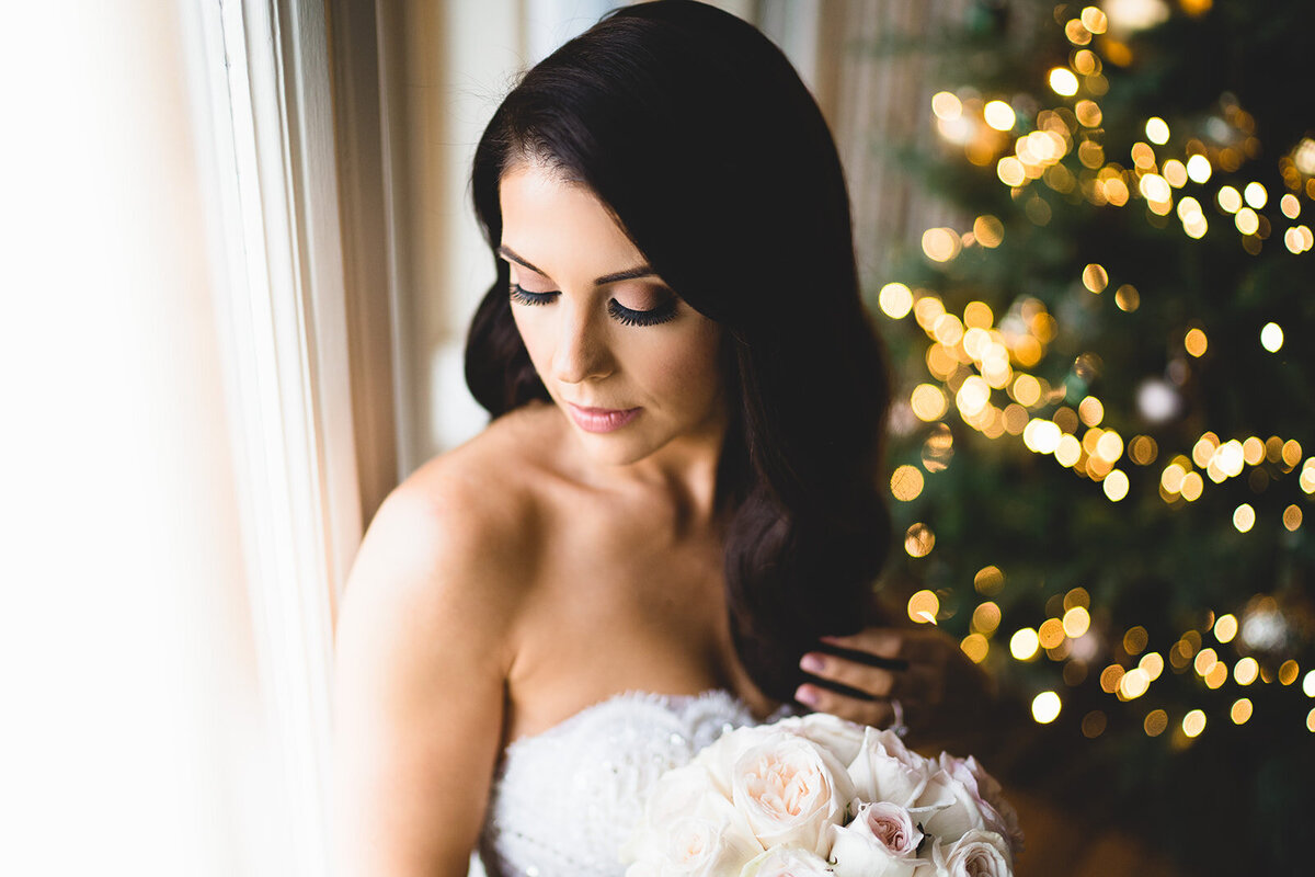 The bride sat by the window looking down holding her flowers with wavy hair and Christmas trees in the background