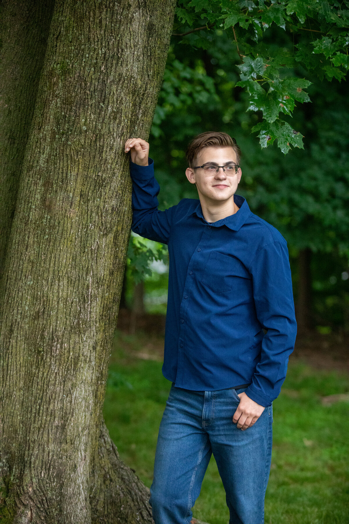 white teenage boy with light brown hair dressed in a blue shirt and blue jeans leans against a tree Clarkstown North senior portrait photographer Milestone Images