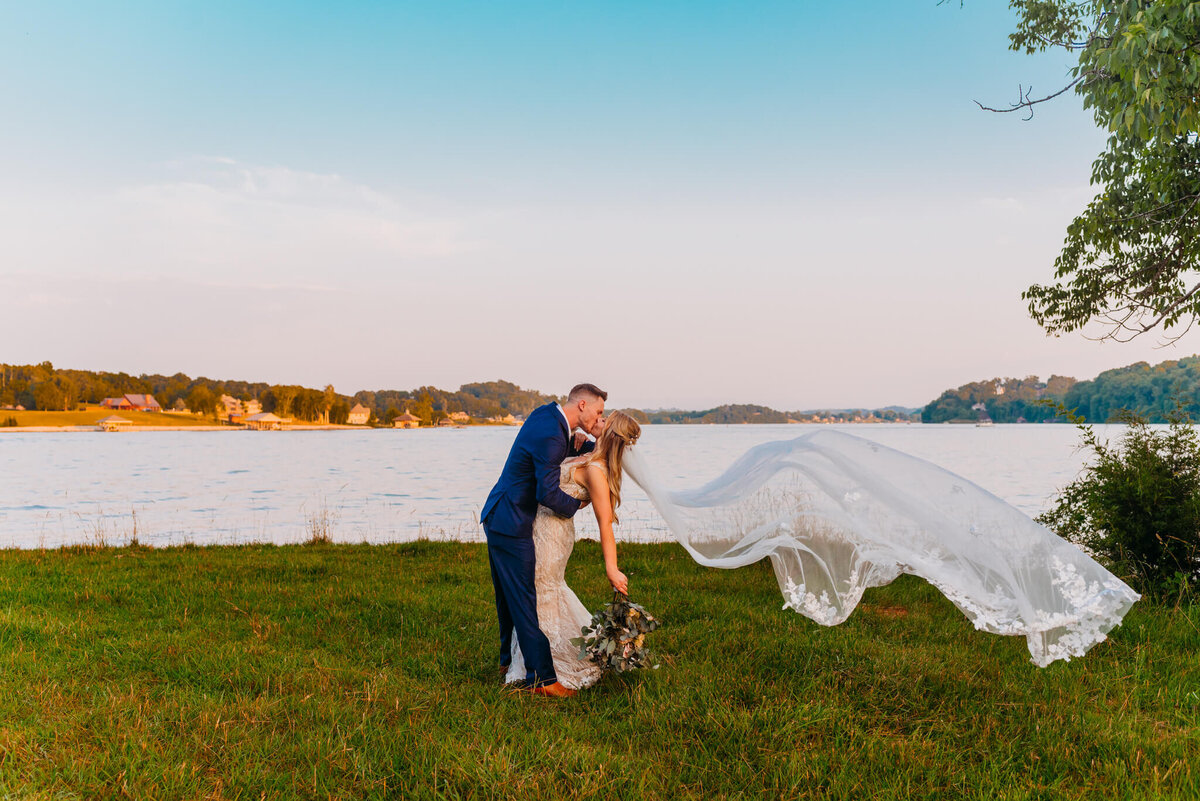 Photo of a bride and groom kissing in front of a river while her veil flows in the wind