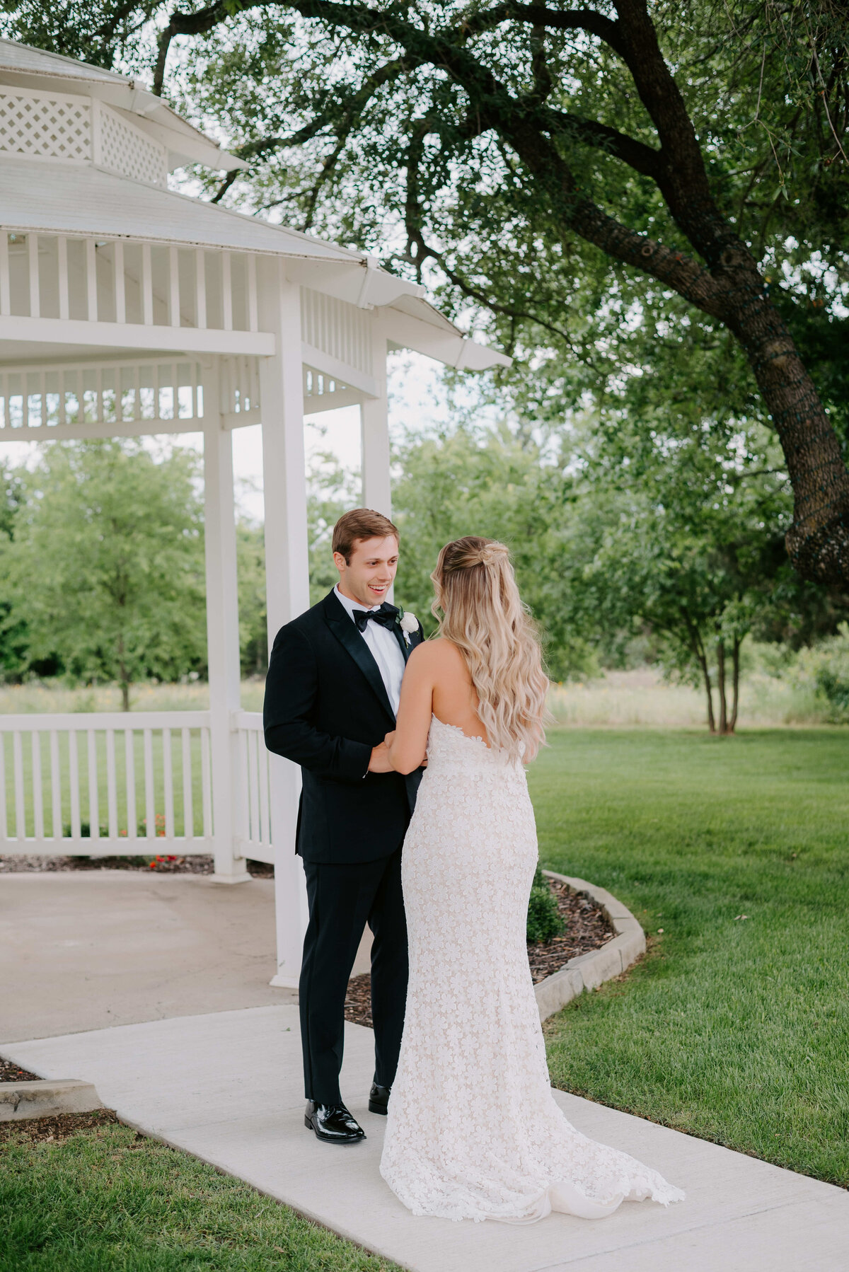 A bride and groom stand near a white gazebo surrounded by lush greenery. The bride wears a white gown, and the groom is in a black tuxedo, creating an elegant and romantic atmosphere.