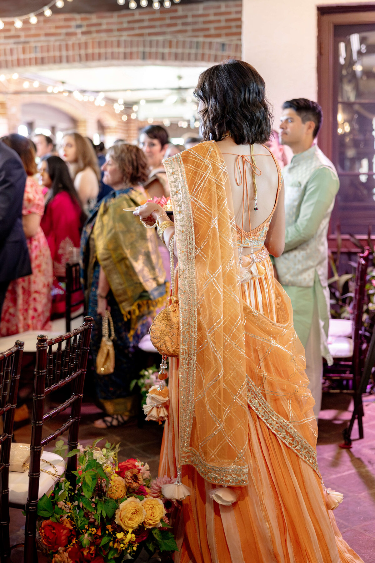 A person in a vibrant, orange traditional attire stands in a beautifully decorated indoor setting, holding a flower bouquet. They are facing away, surrounded by seated guests, some in colorful clothing, with lights hanging overhead.