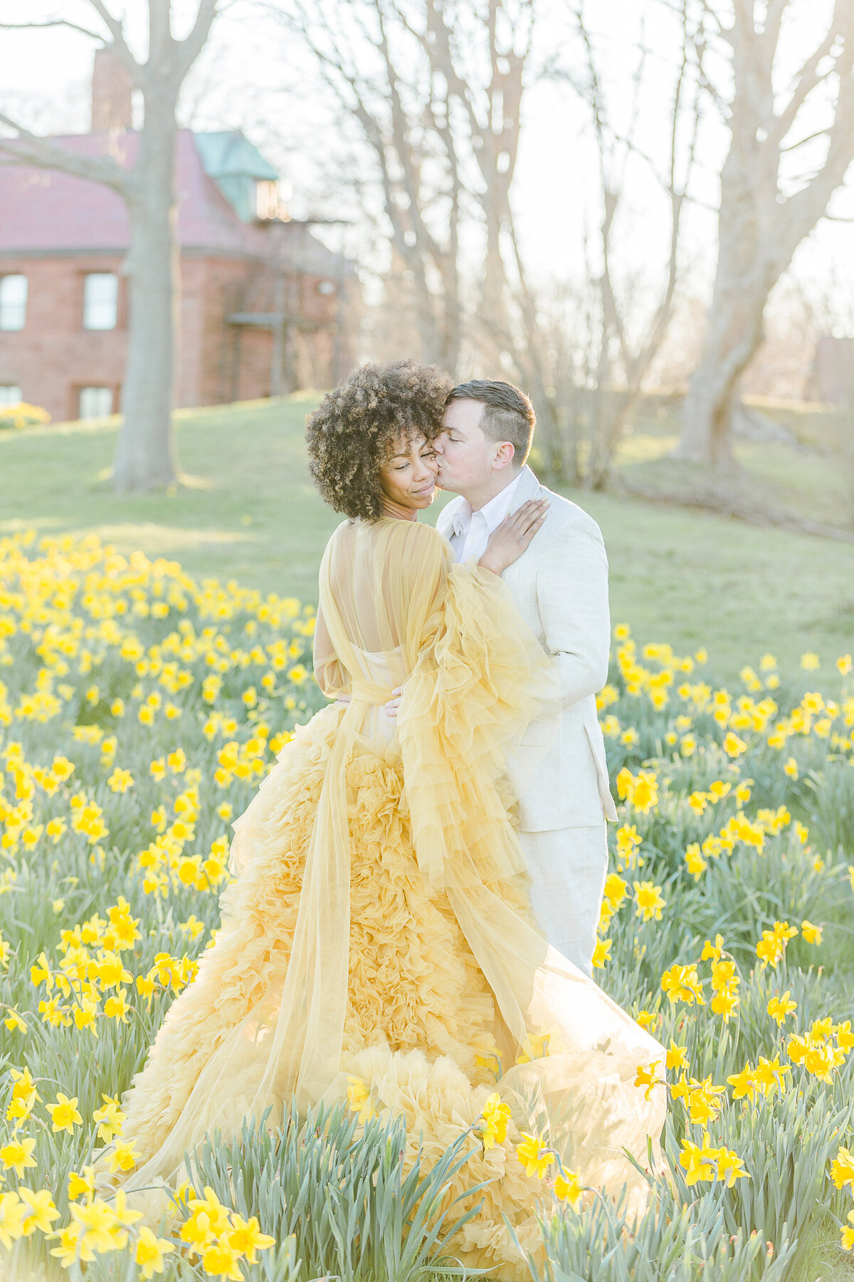 Salve Regina Cliff Walk engagement photoshoot. Man and woman are standing facing each other amongst daffodils. The woman is in a long yellow organza gown with her hands on her fiance's chest. The man is gently kissing the woman on the cheek. Captured by best RI wedding and engagement photographer Lia Rose Weddings